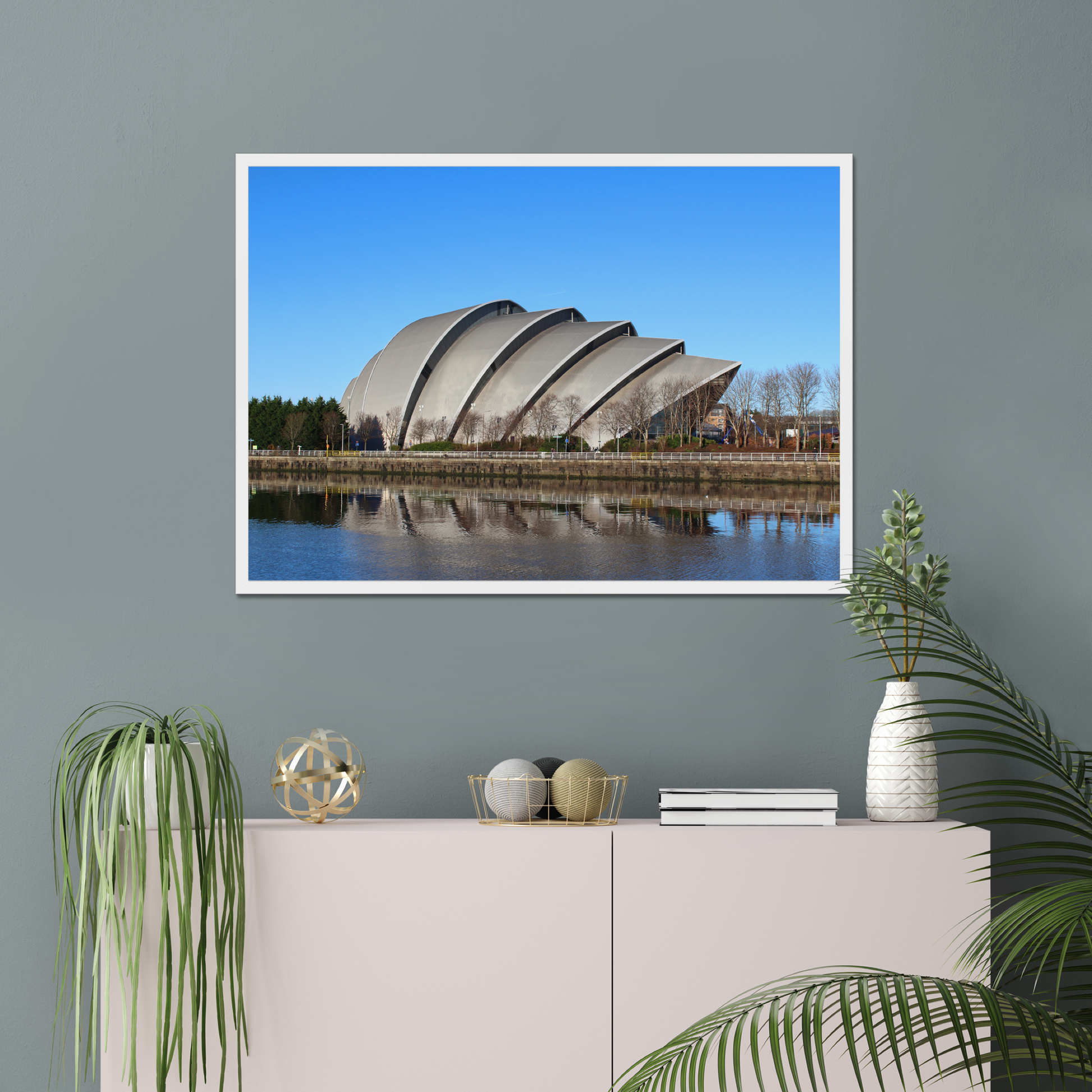 Modern architectural building reflected in water with a clear blue sky on a gray wall above a white cabinet with decorative items.