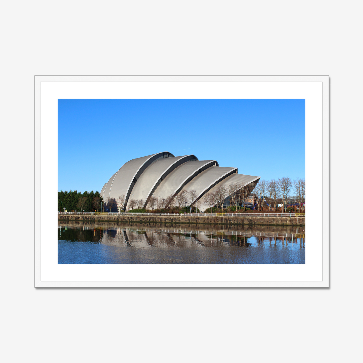 Framed photograph of a modern building with a curved roof, reflected in water, against a blue sky.