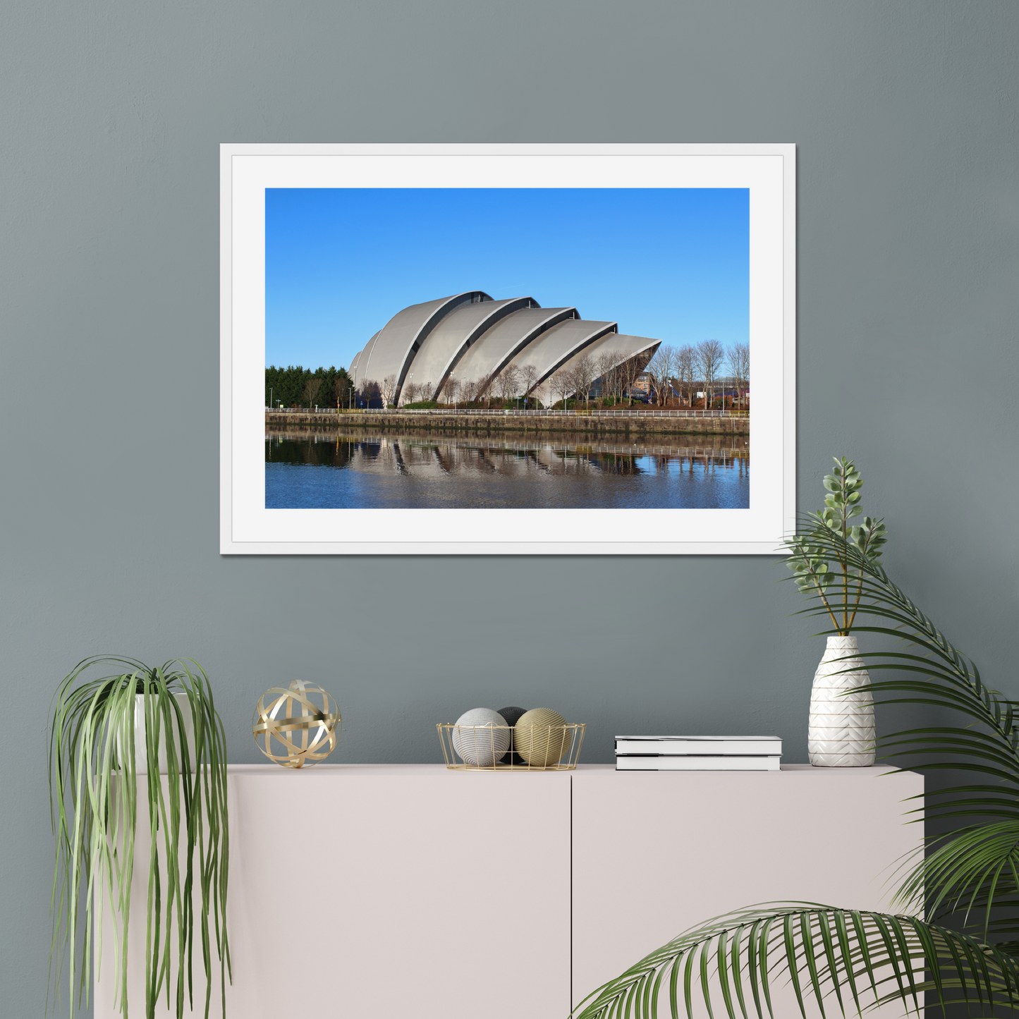 Modern architectural building reflected in water with a clear blue sky on a gray wall above a white cabinet with decorative items.