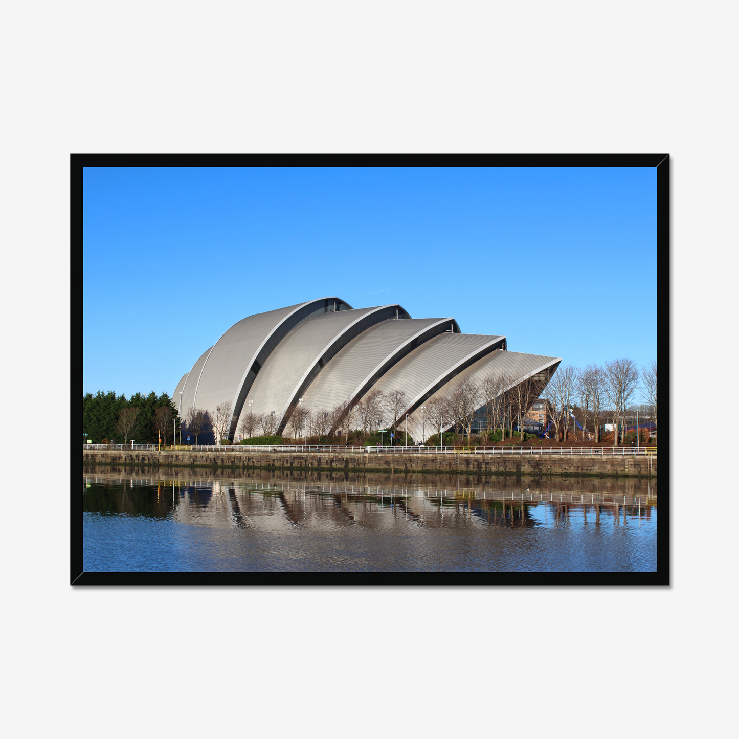 Framed photograph of a modern building with a curved roof, reflected in water, against a blue sky.