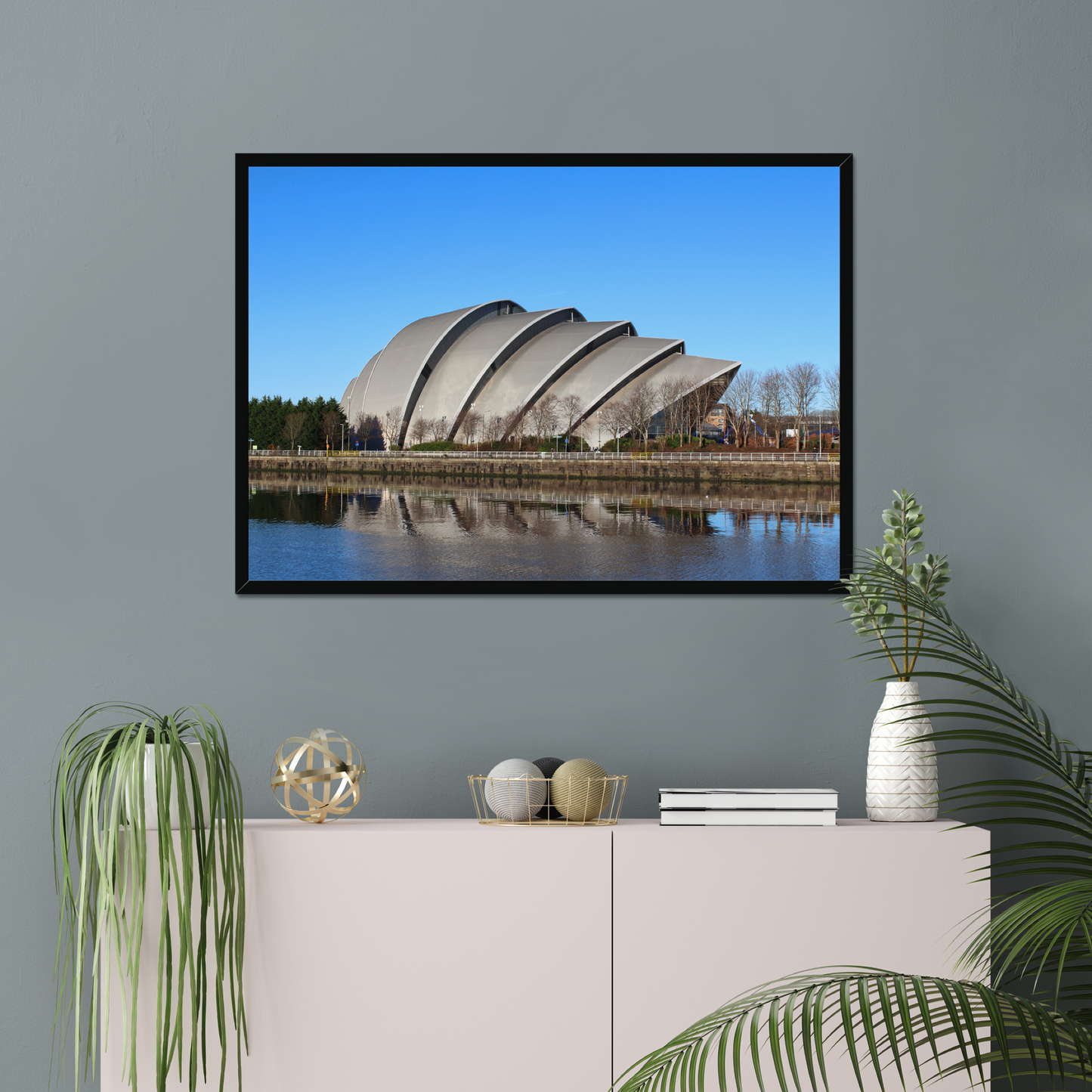 Modern architectural building reflected in water with a clear blue sky on a gray wall above a white cabinet with decorative items.
