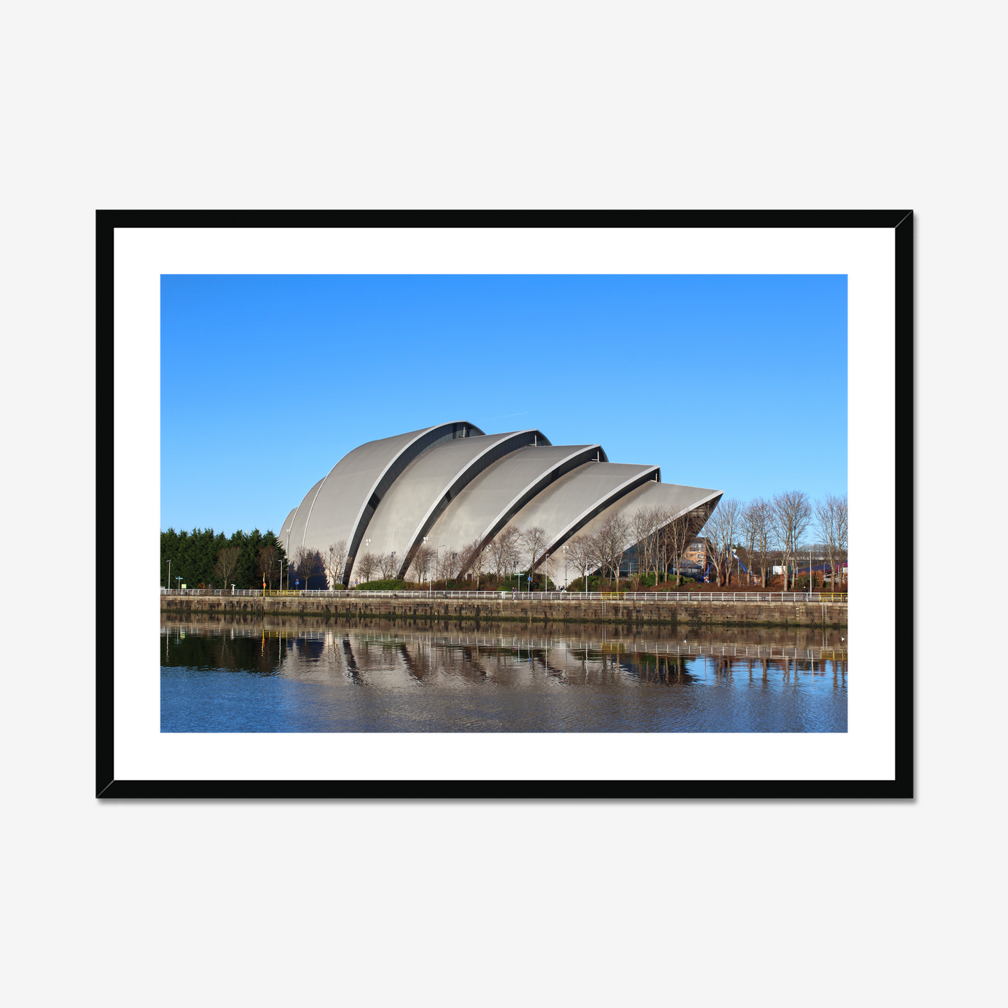 Framed photograph of a modern building with a curved roof, reflected in water, against a blue sky.