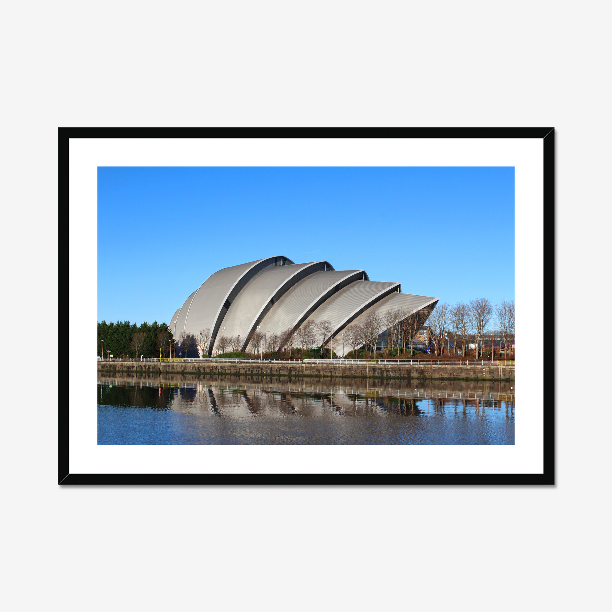 Framed photograph of a modern building with a curved roof, reflected in water, against a blue sky.