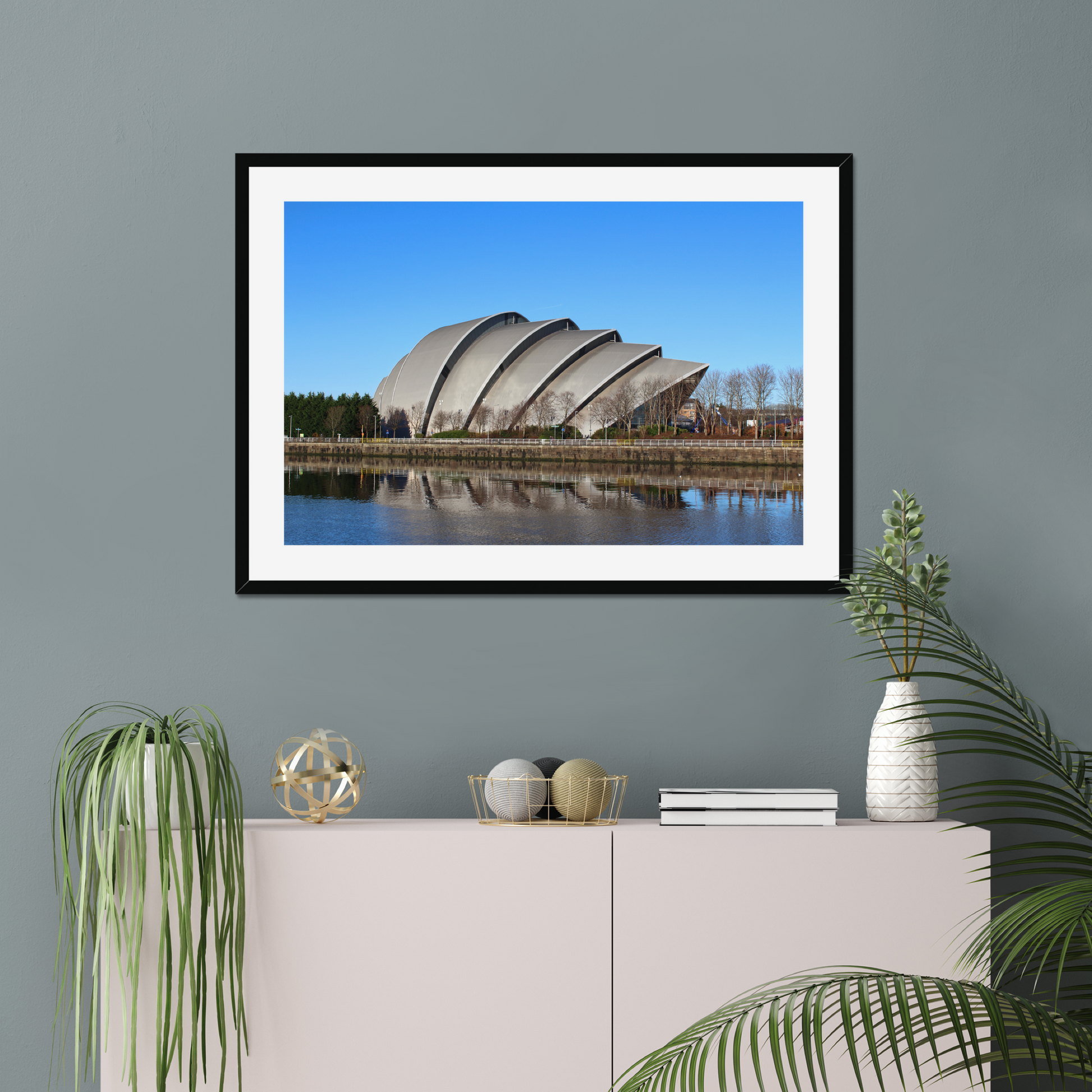 Modern architectural building reflected in water with a clear blue sky on a gray wall above a white cabinet with decorative items.