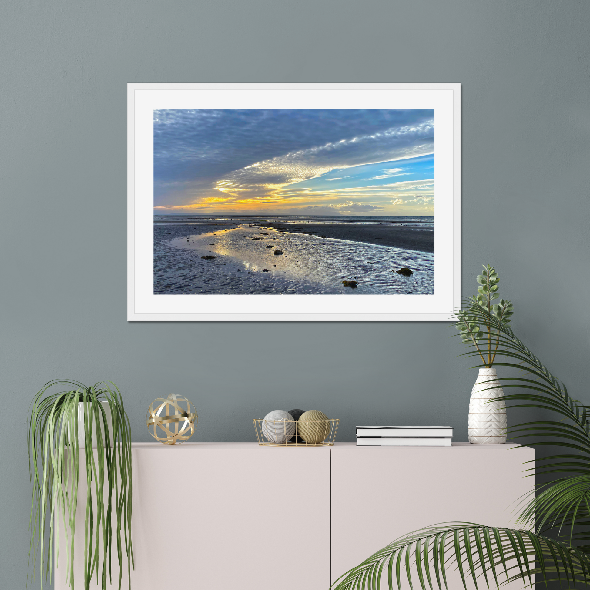 Framed photograph of a sunset over a beach with clouds and water reflections on a wall above a cabinet with decorative items.