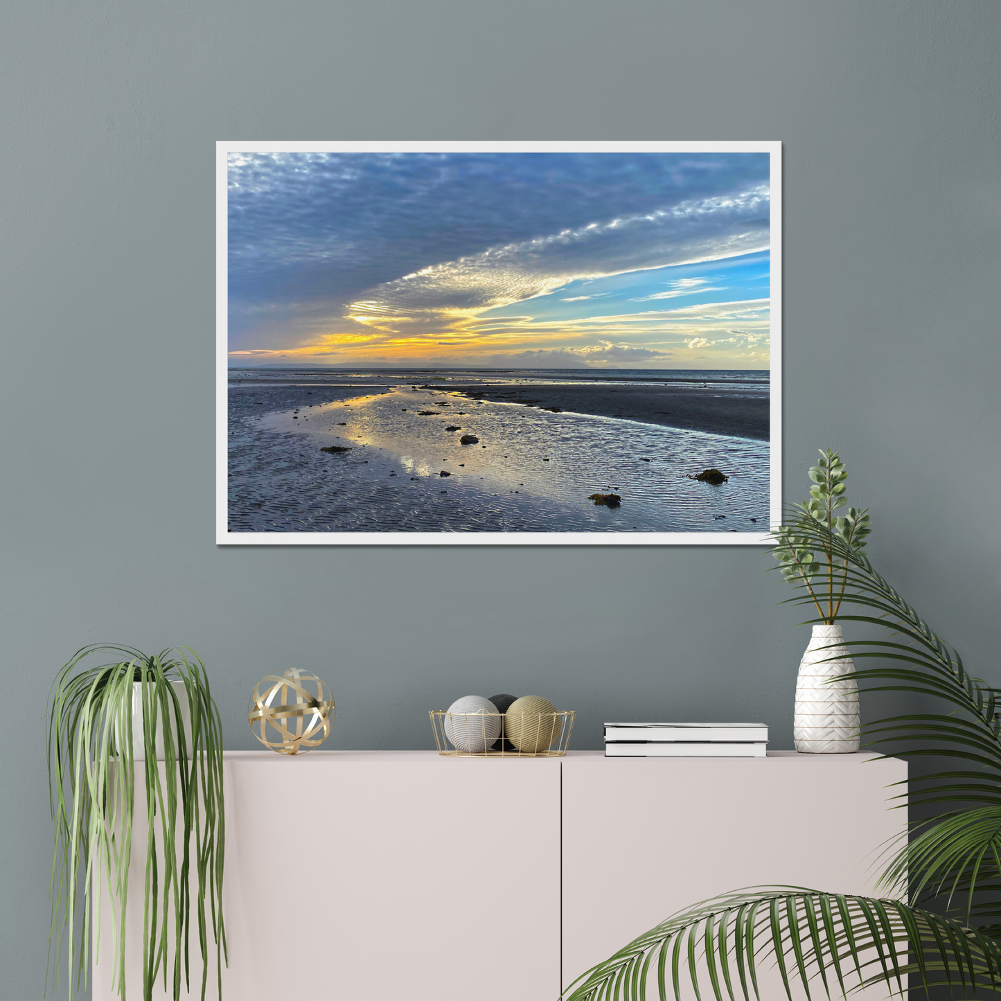 Framed photograph of a sunset over a beach with clouds and water reflections on a wall above a cabinet with decorative items.