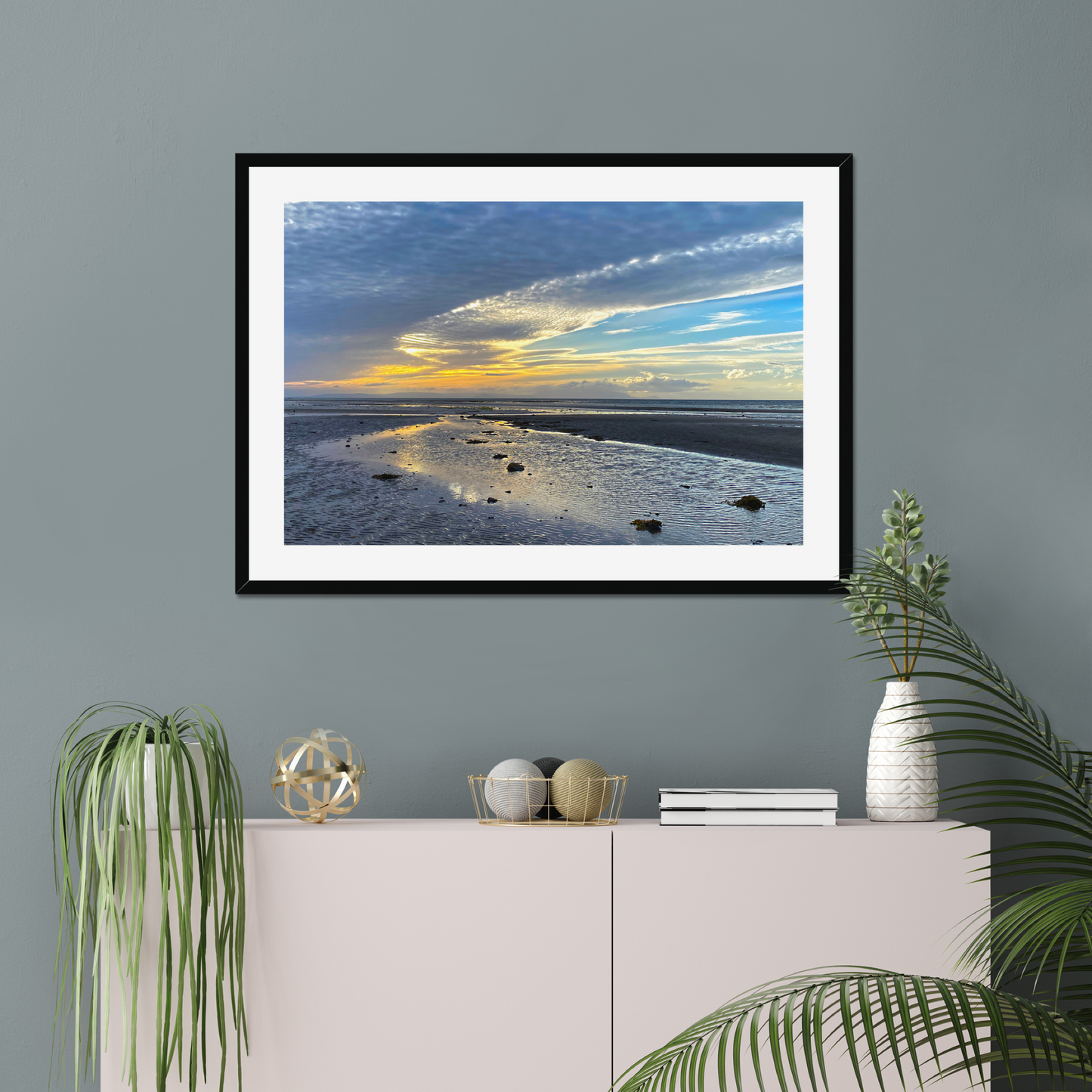 Framed photograph of a sunset over a beach with clouds and water reflections on a wall above a cabinet with decorative items.