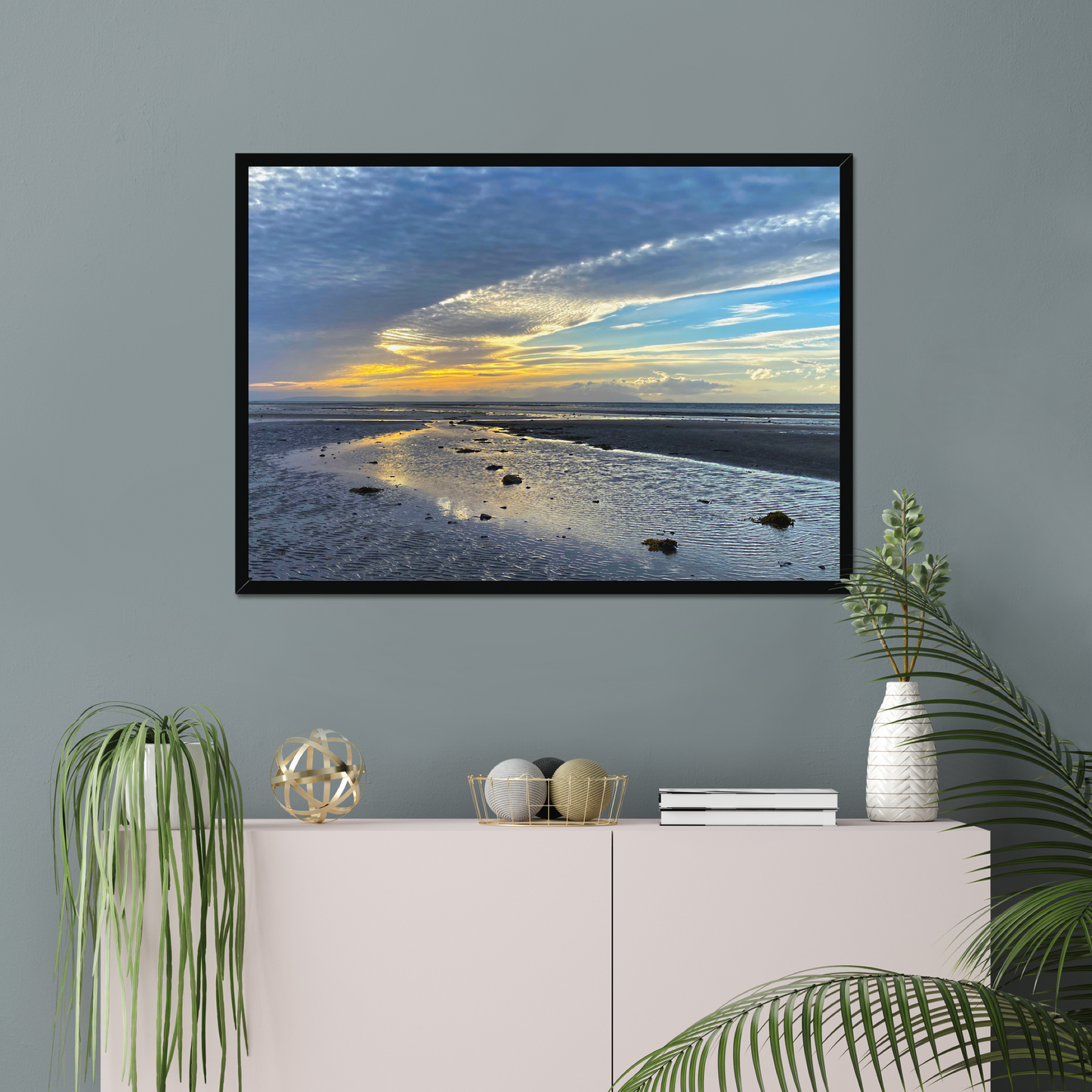 Framed photograph of a sunset over a beach with clouds and water reflections on a wall above a cabinet with decorative items.