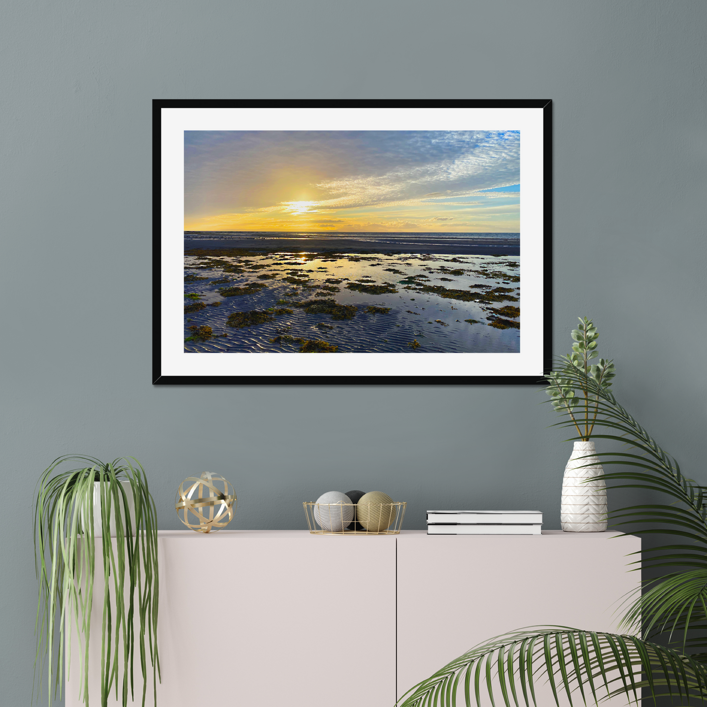 Framed artwork of a sunset over water on a gray wall above a white cabinet with decorative items.