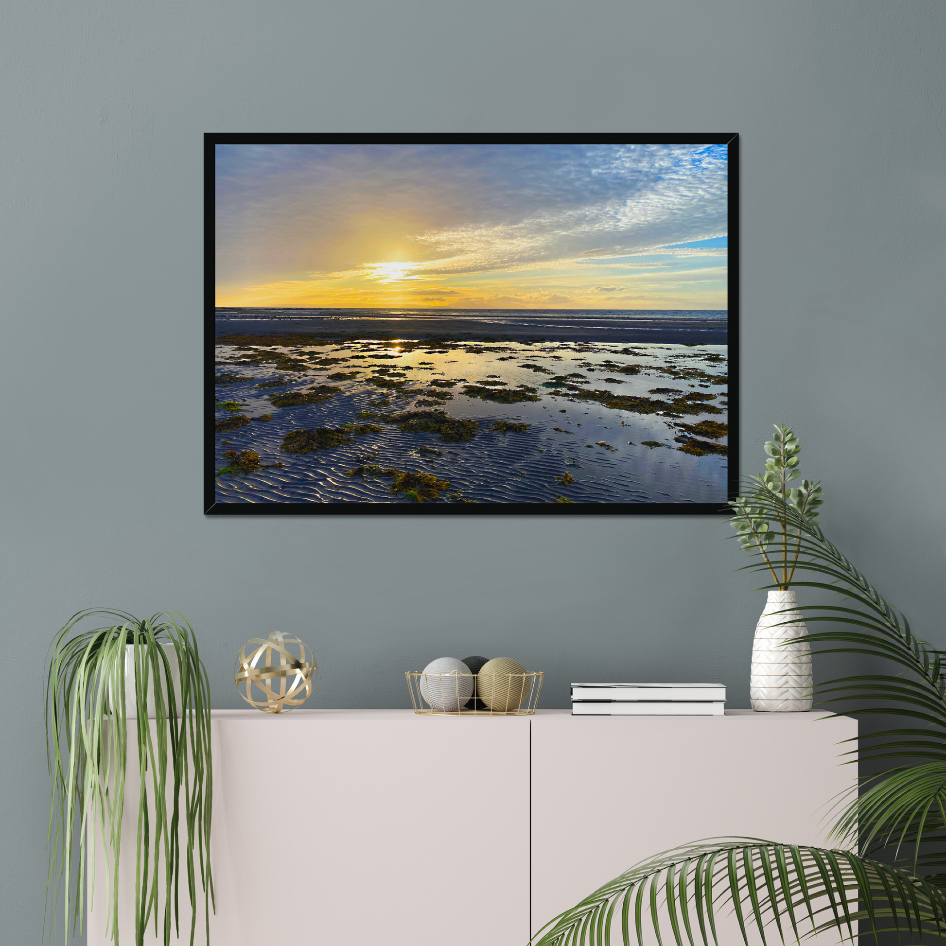 Framed artwork of a sunset over water on a gray wall above a white console table with decorative items.