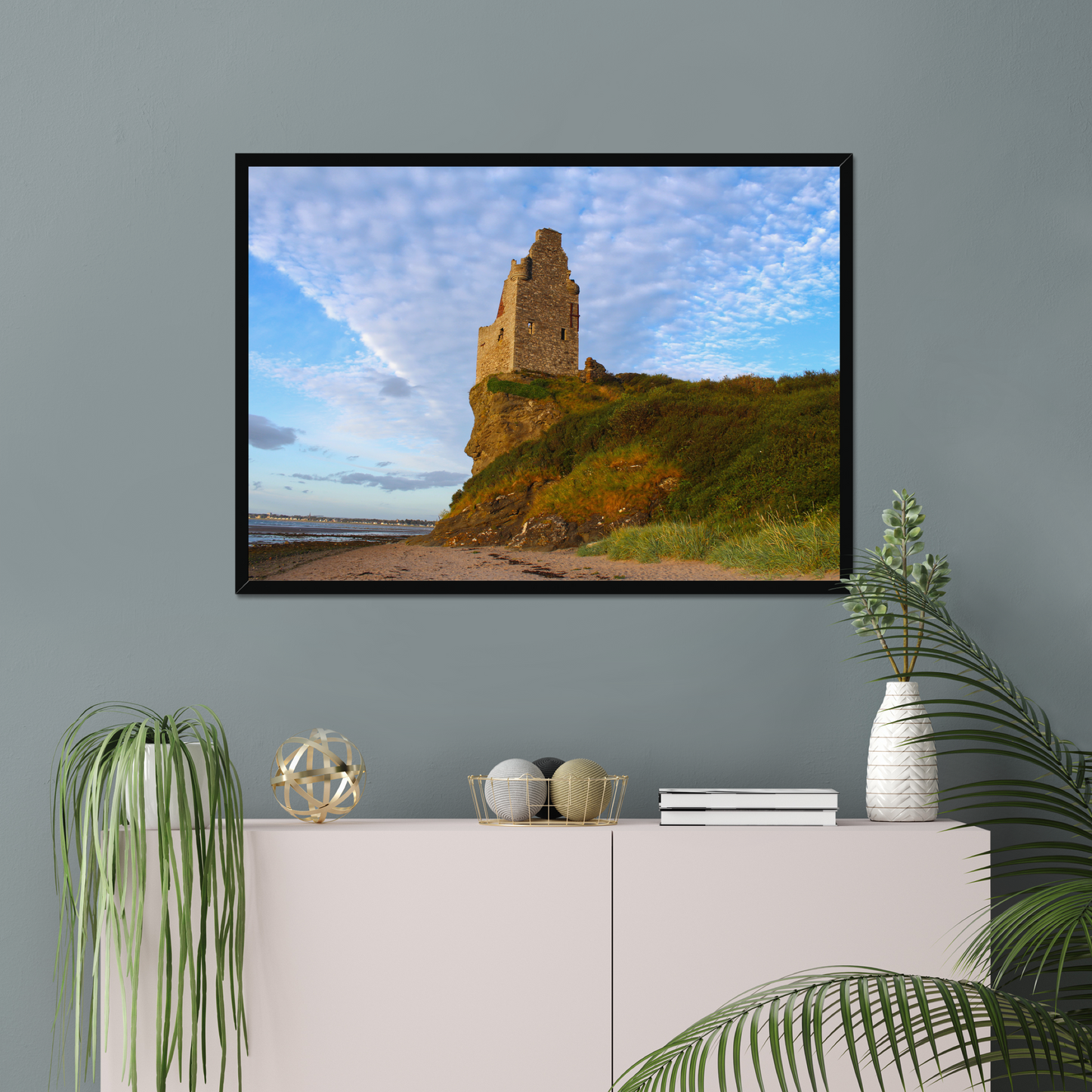 Framed picture of a coastal scene with a castle on a wall above a white cabinet with decorative items.