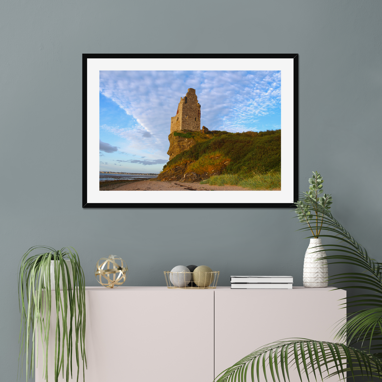Framed picture of a coastal scene with a castle on a wall above a white cabinet with decorative items.