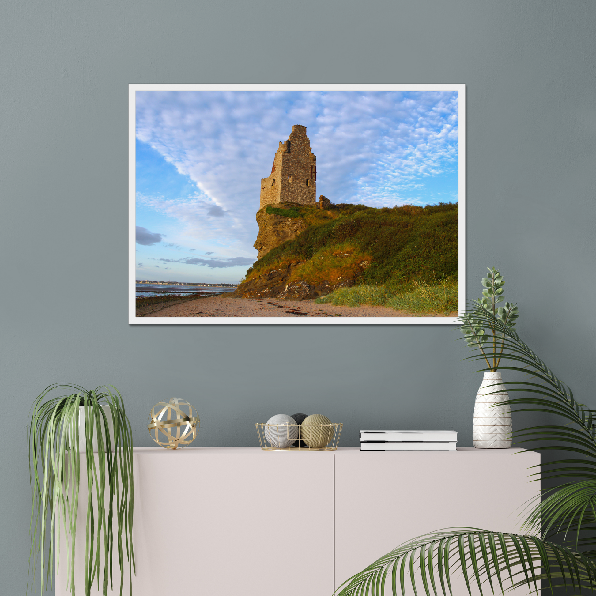 Framed picture of a coastal scene with a castle on a wall above a white cabinet with decorative items.