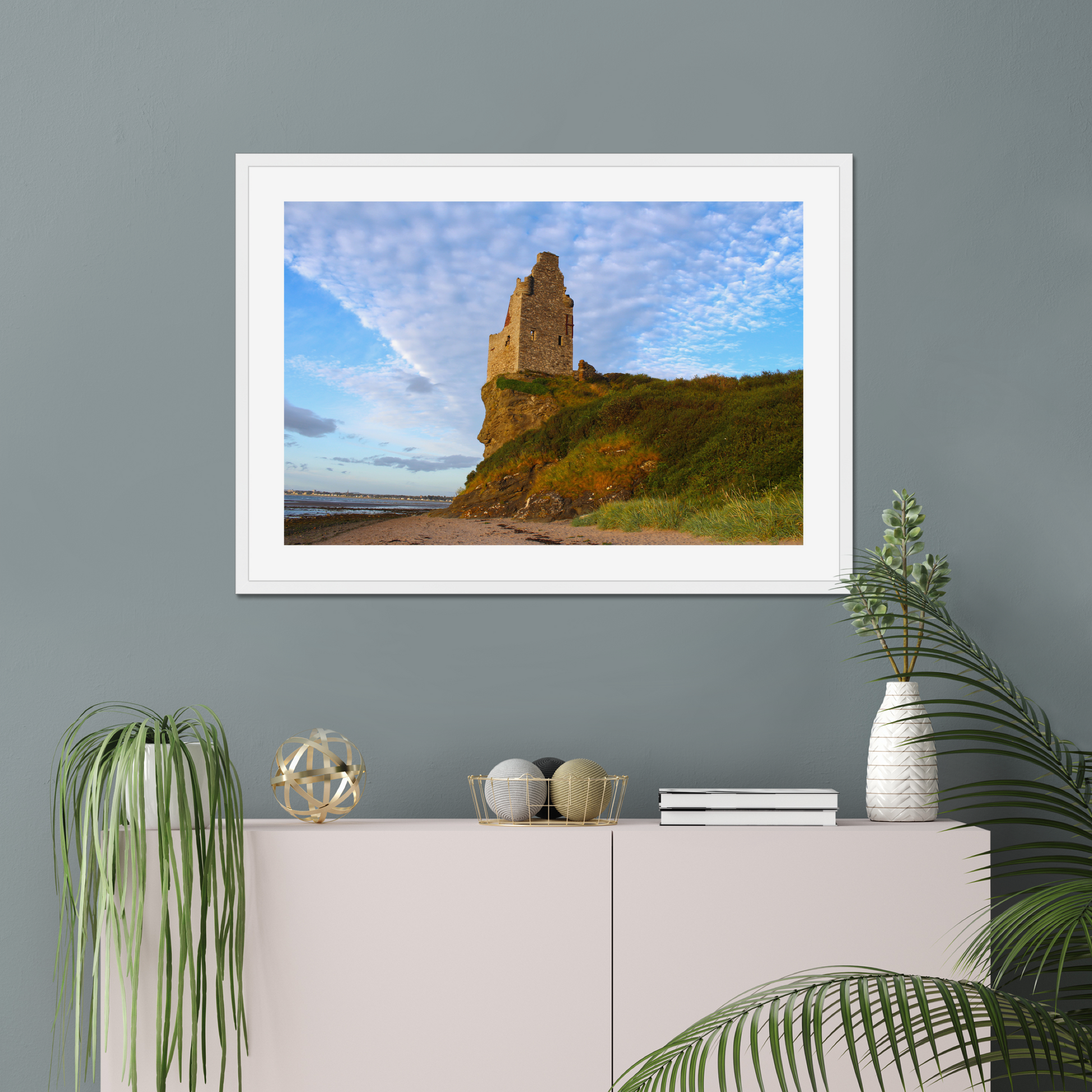 Framed picture of a coastal scene with a castle on a wall above a white cabinet with decorative items.