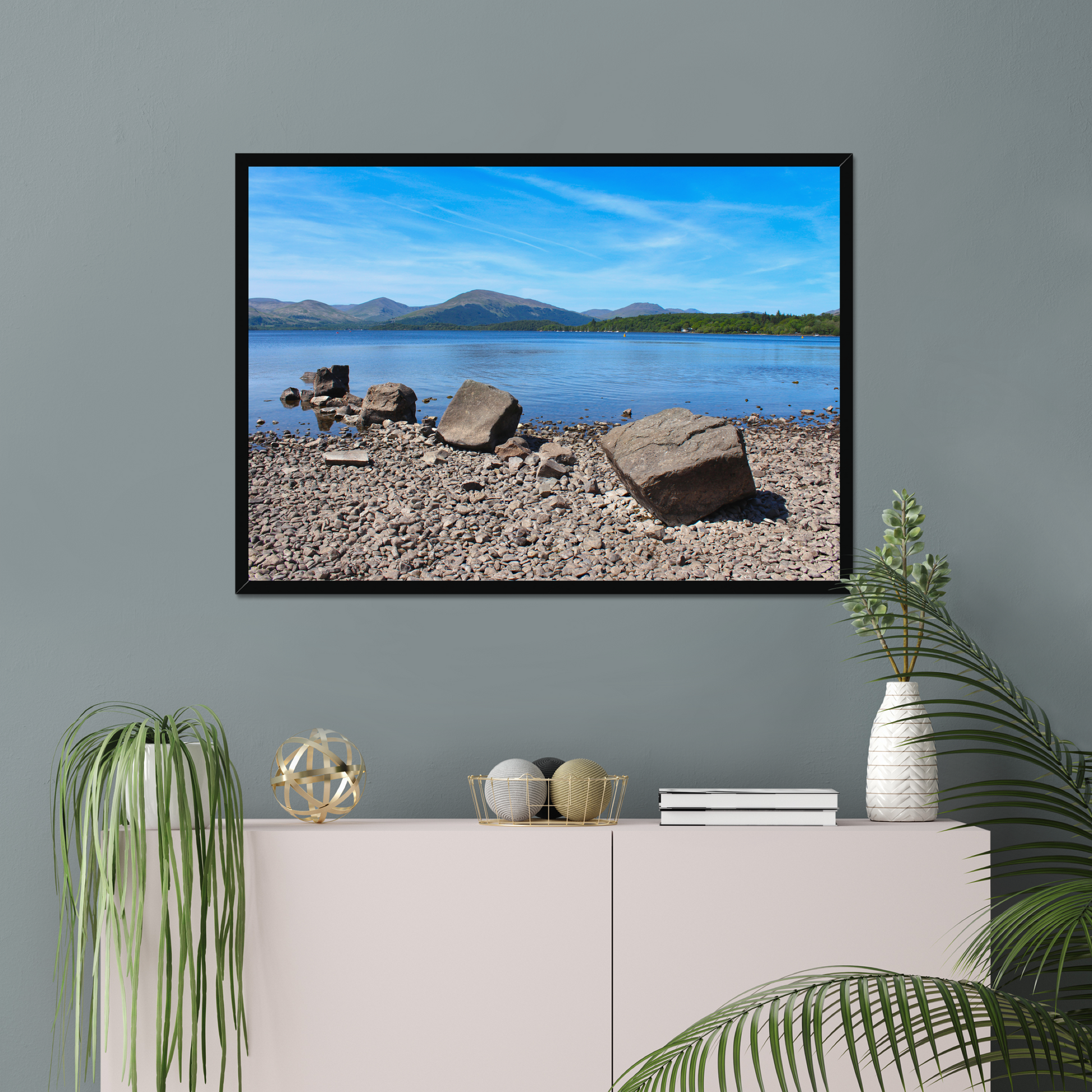 Framed picture of a lake with mountains on a gray wall above a white cabinet with decorative items.