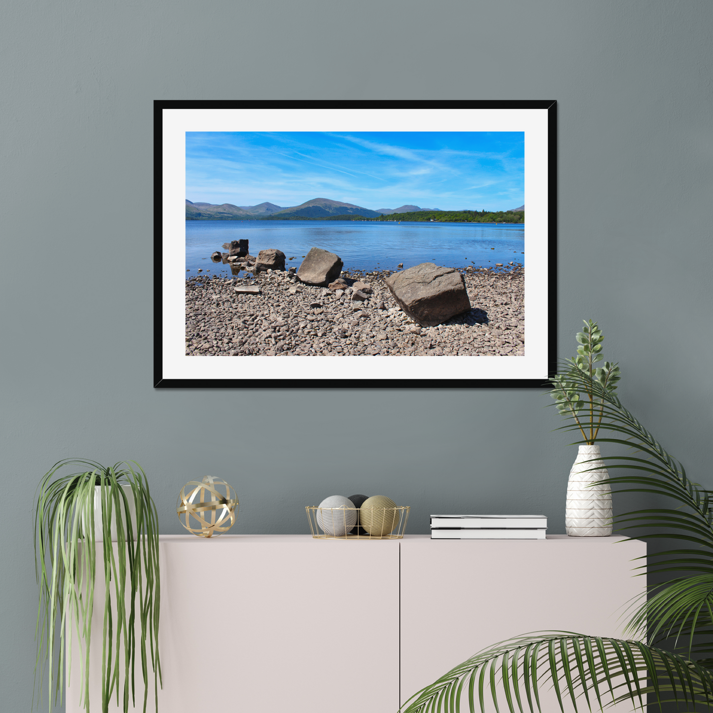 Framed landscape print of a lake and mountains on a gray wall above a white cabinet with decorative items.