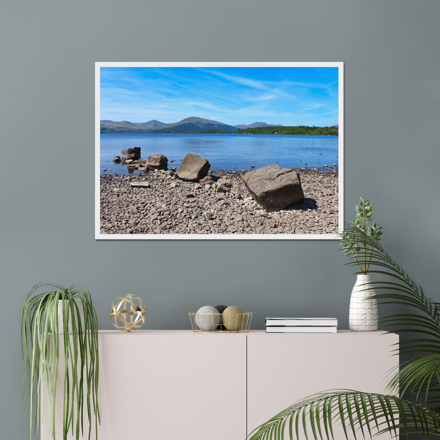 Framed landscape print of a lake and mountains on a gray wall above a white cabinet with decorative items.