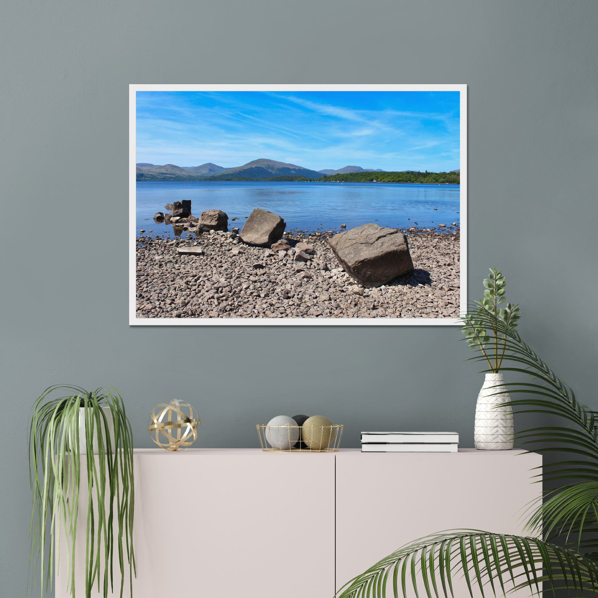 Framed landscape print of a lake and mountains on a gray wall above a white cabinet with decorative items.