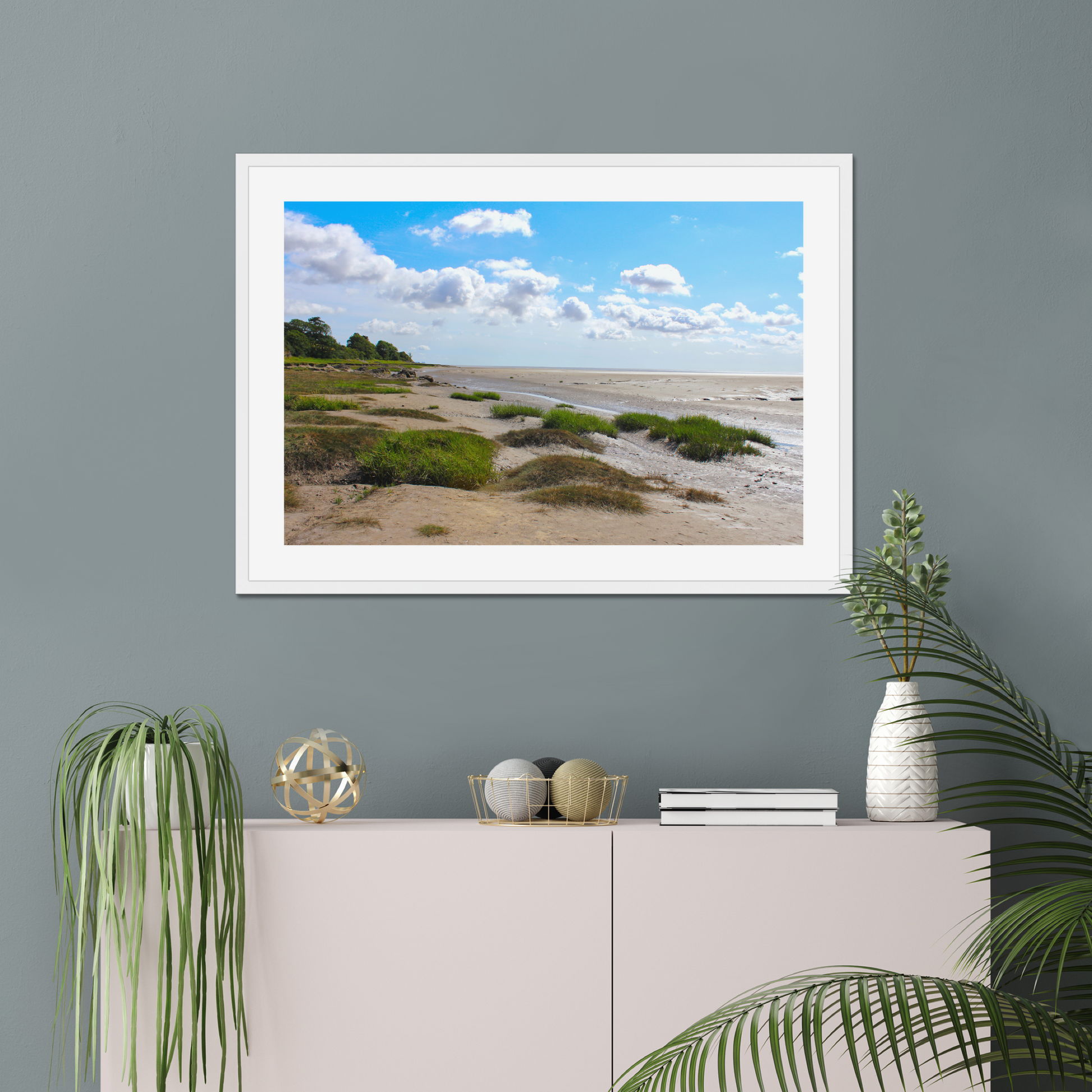 Framed landscape print of a beach scene on a gray wall above a white cabinet with decorative items.