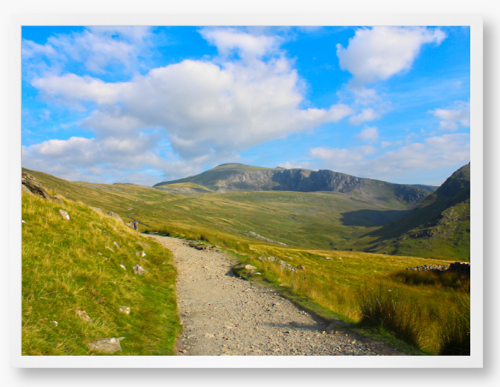 Framed photo of a mountain landscape with a path leading towards distant mountains under a blue sky with clouds