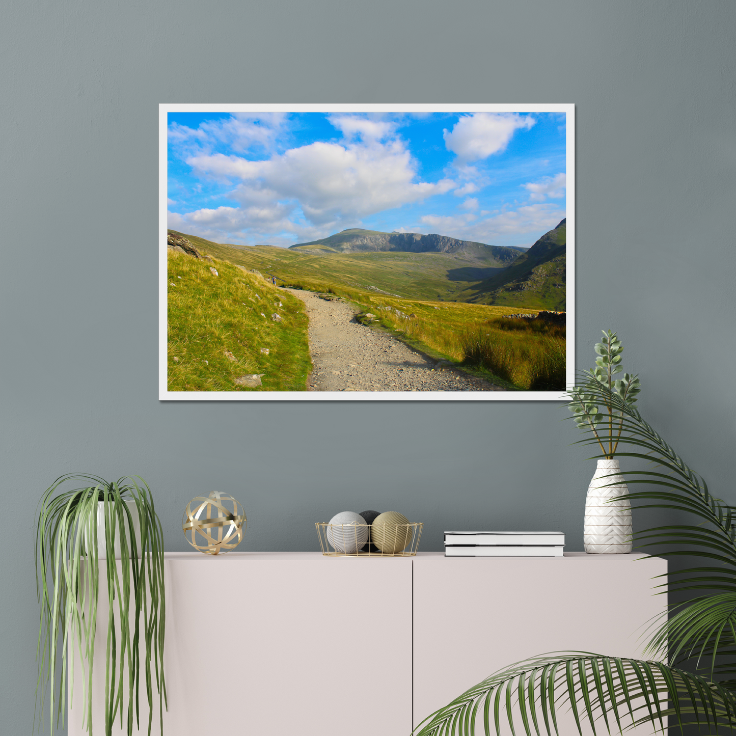Framed photograph of a mountain landscape with a clear blue sky and a path leading towards the mountains on a gray wall above a white console table with decorative items.
