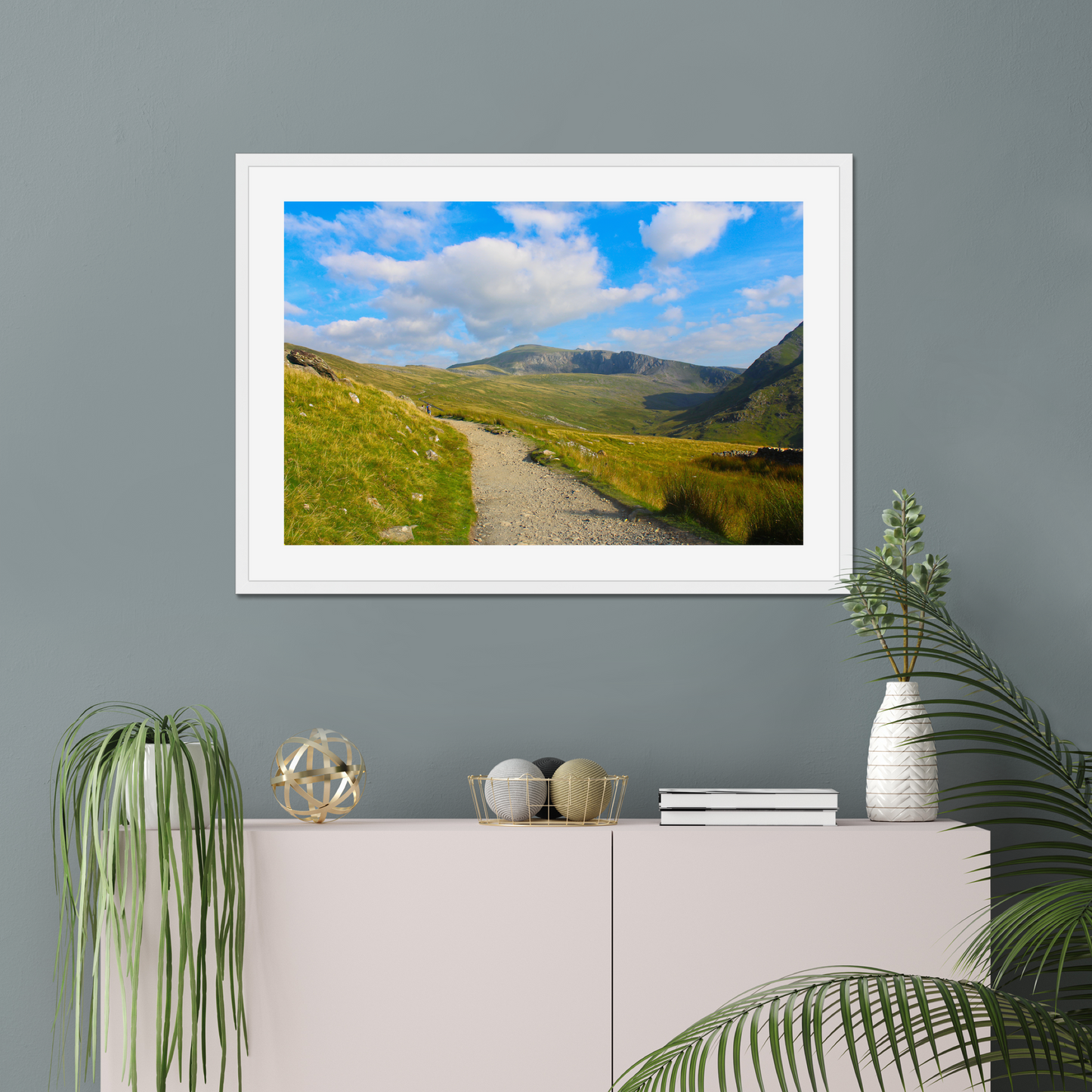 Framed photograph of a mountain landscape with a clear blue sky and a path leading towards the mountains on a gray wall above a white console table with decorative items.