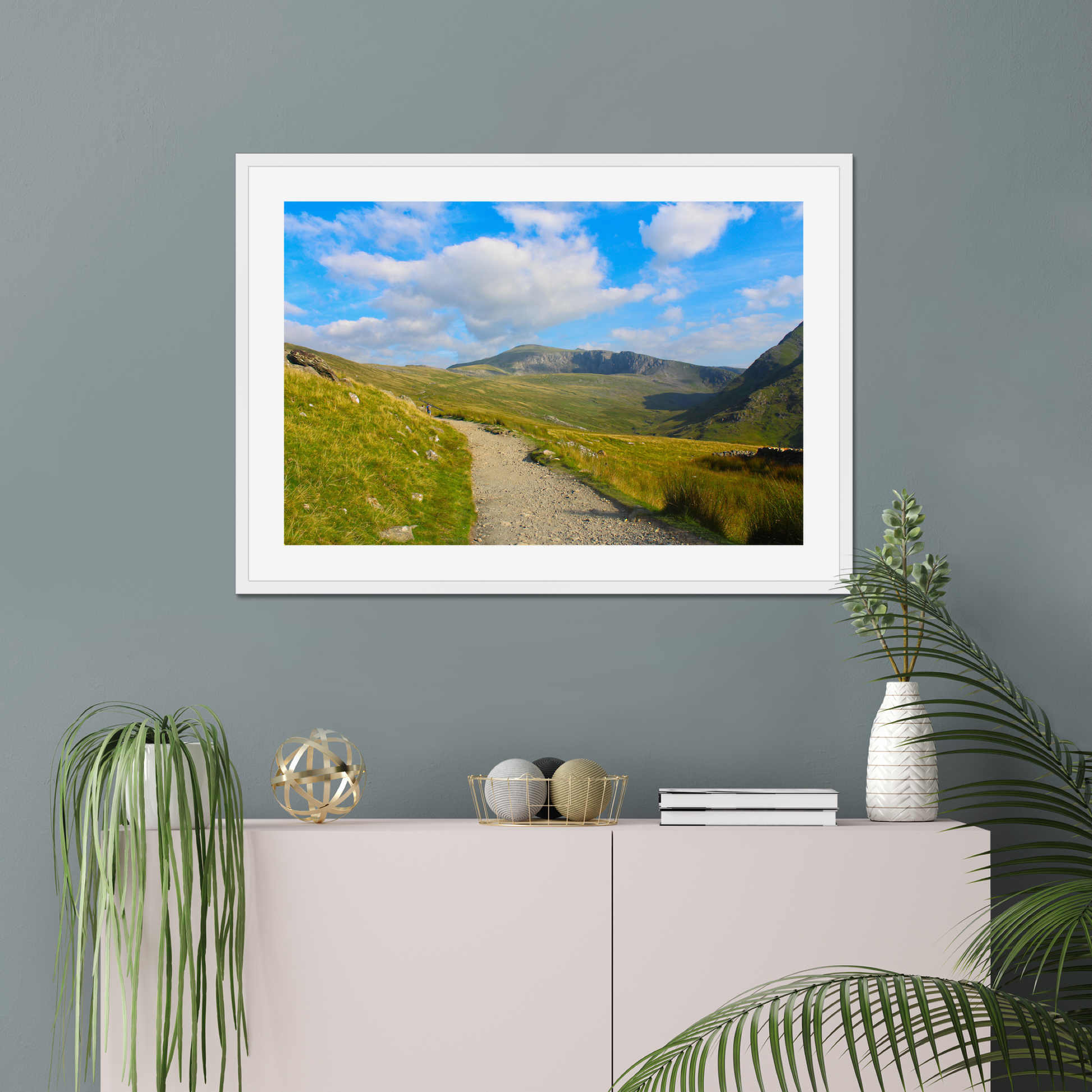Framed photograph of a mountain landscape with a clear blue sky and a path leading towards the mountains on a gray wall above a white console table with decorative items.
