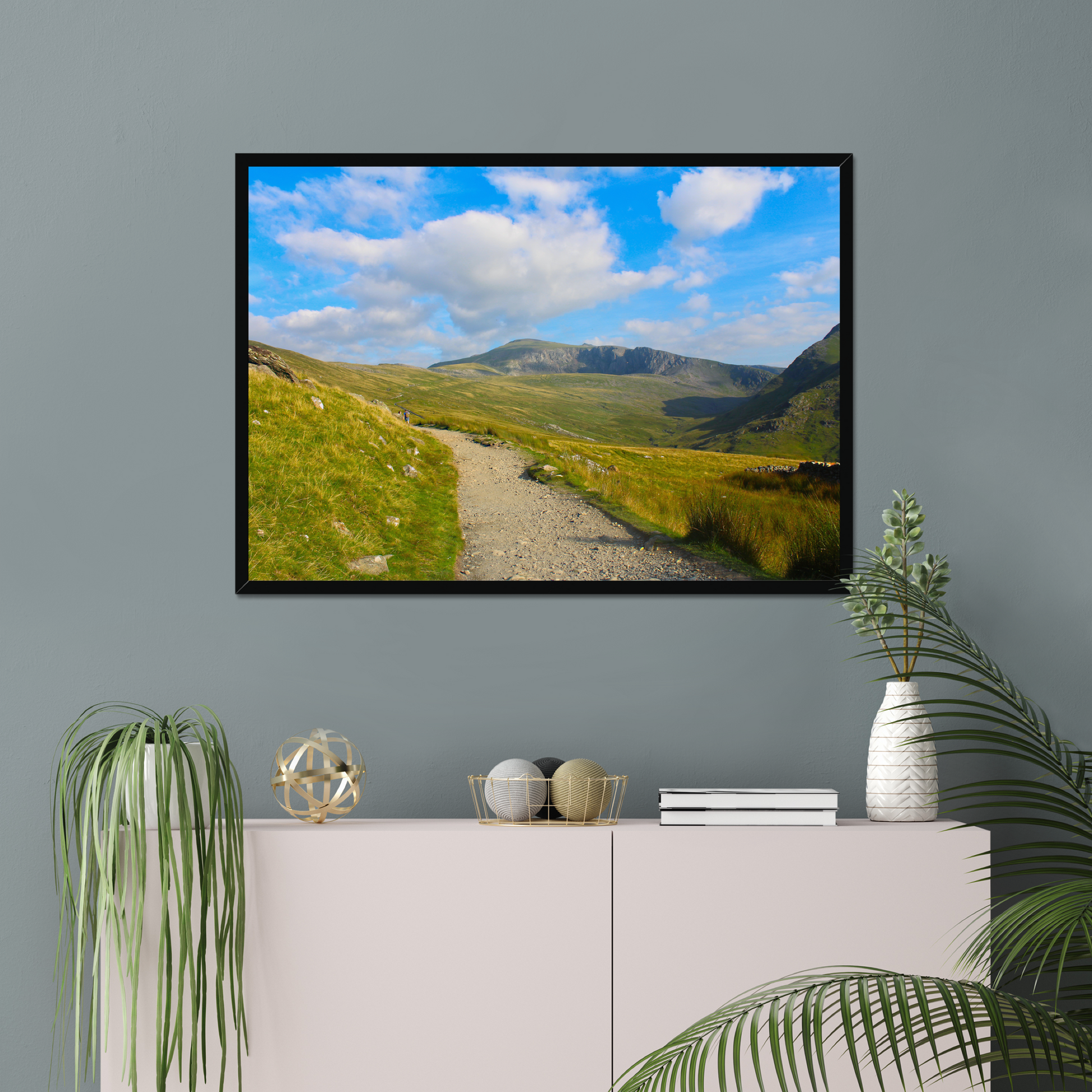 Framed photograph of a mountain landscape with a clear blue sky and a path leading towards the mountains on a gray wall above a white console table with decorative items.