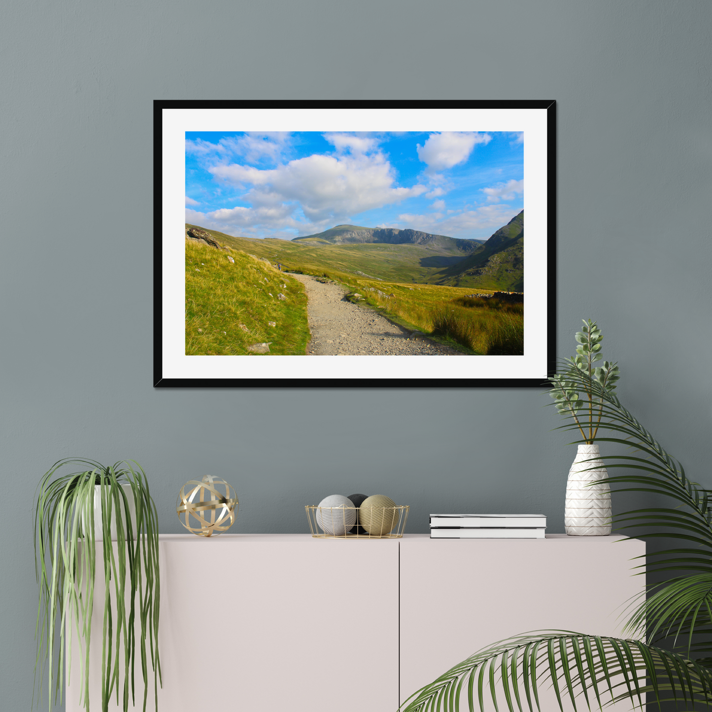 Framed photograph of a mountain landscape with a clear blue sky and a path leading towards the mountains on a gray wall above a white console table with decorative items.