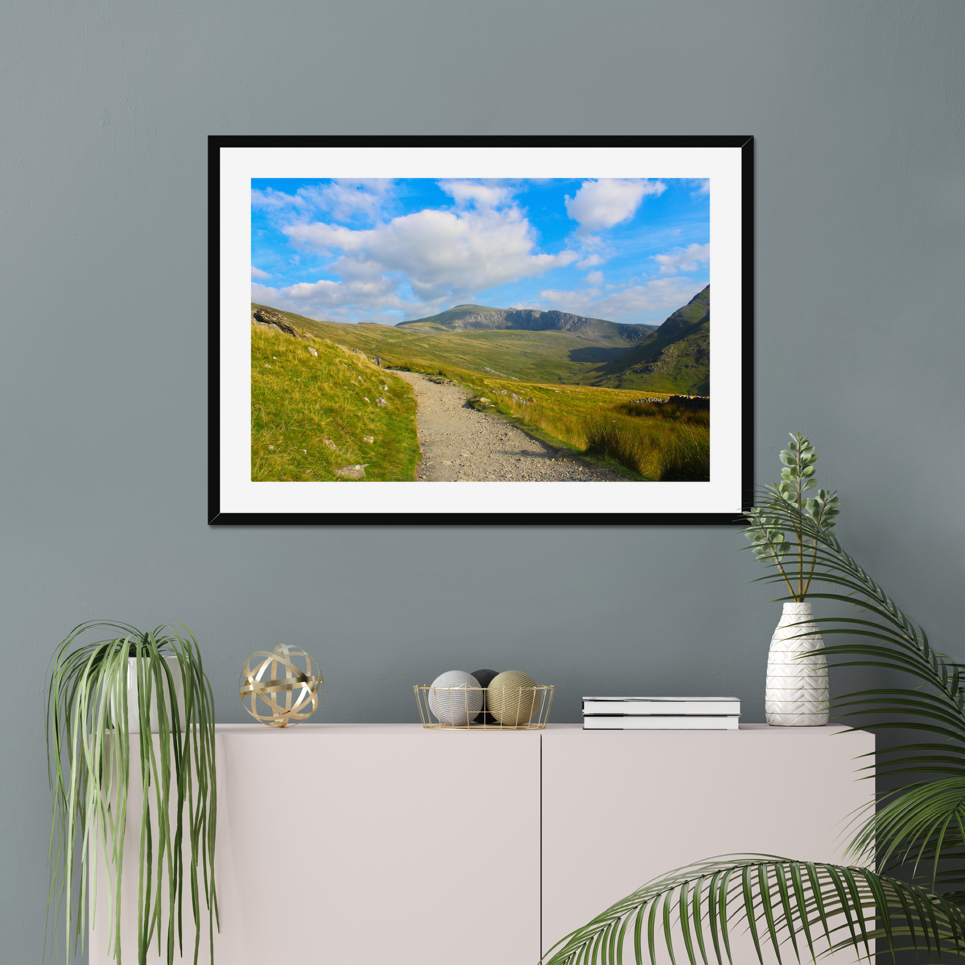 Framed photograph of a mountain landscape with a clear blue sky and a path leading towards the mountains on a gray wall above a white console table with decorative items.