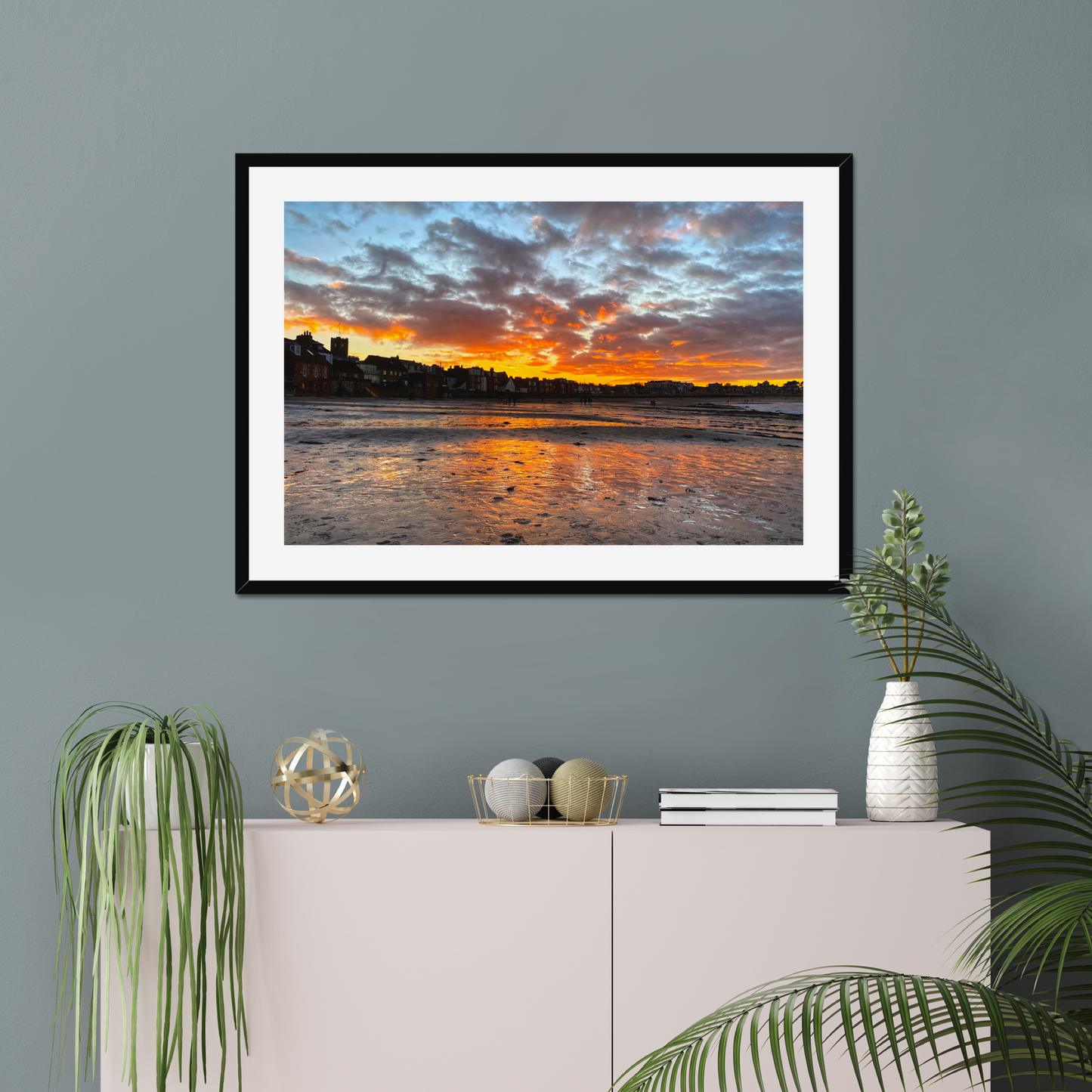 Framed artwork of a sunset over water on a gray wall above a white cabinet with decorative items.