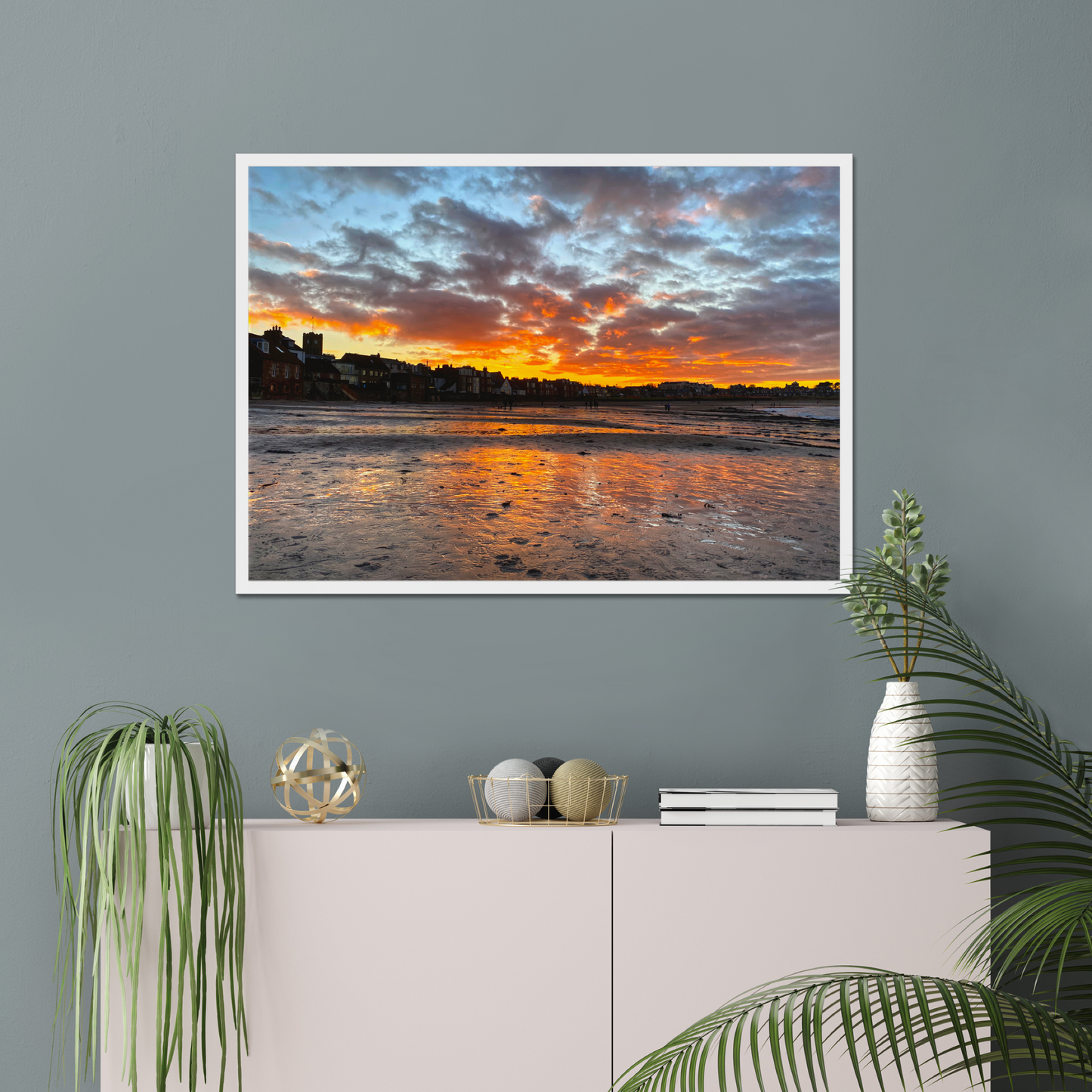 Framed artwork of a sunset over water on a gray wall above a white cabinet with decorative items.