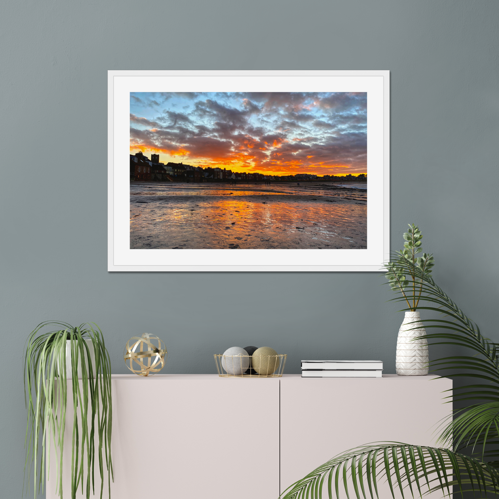 Framed artwork of a sunset over water on a gray wall above a white cabinet with decorative items.