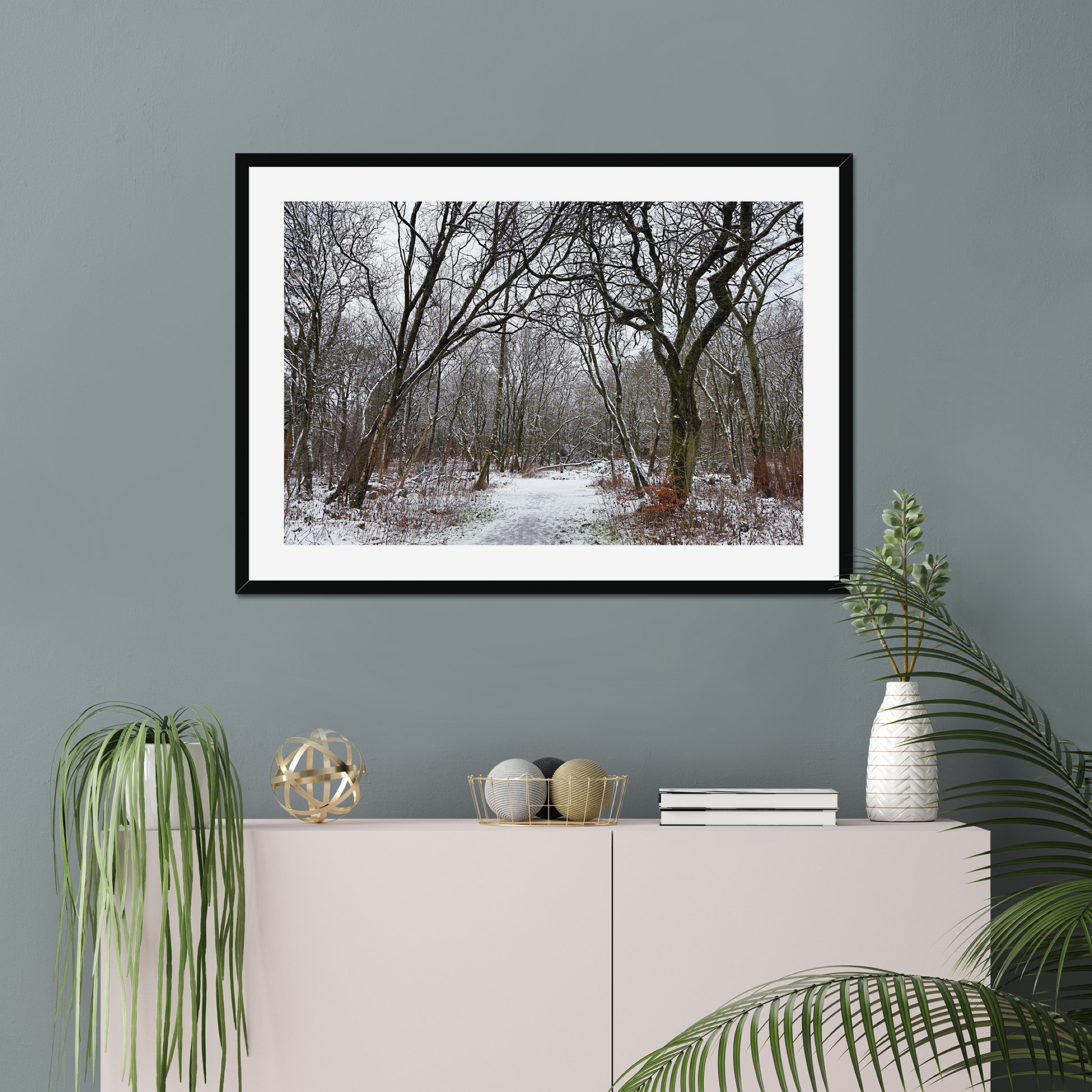 Framed photograph of a winter woodland scene with snow on the ground on a gray wall above a white cabinet with decorative items.
