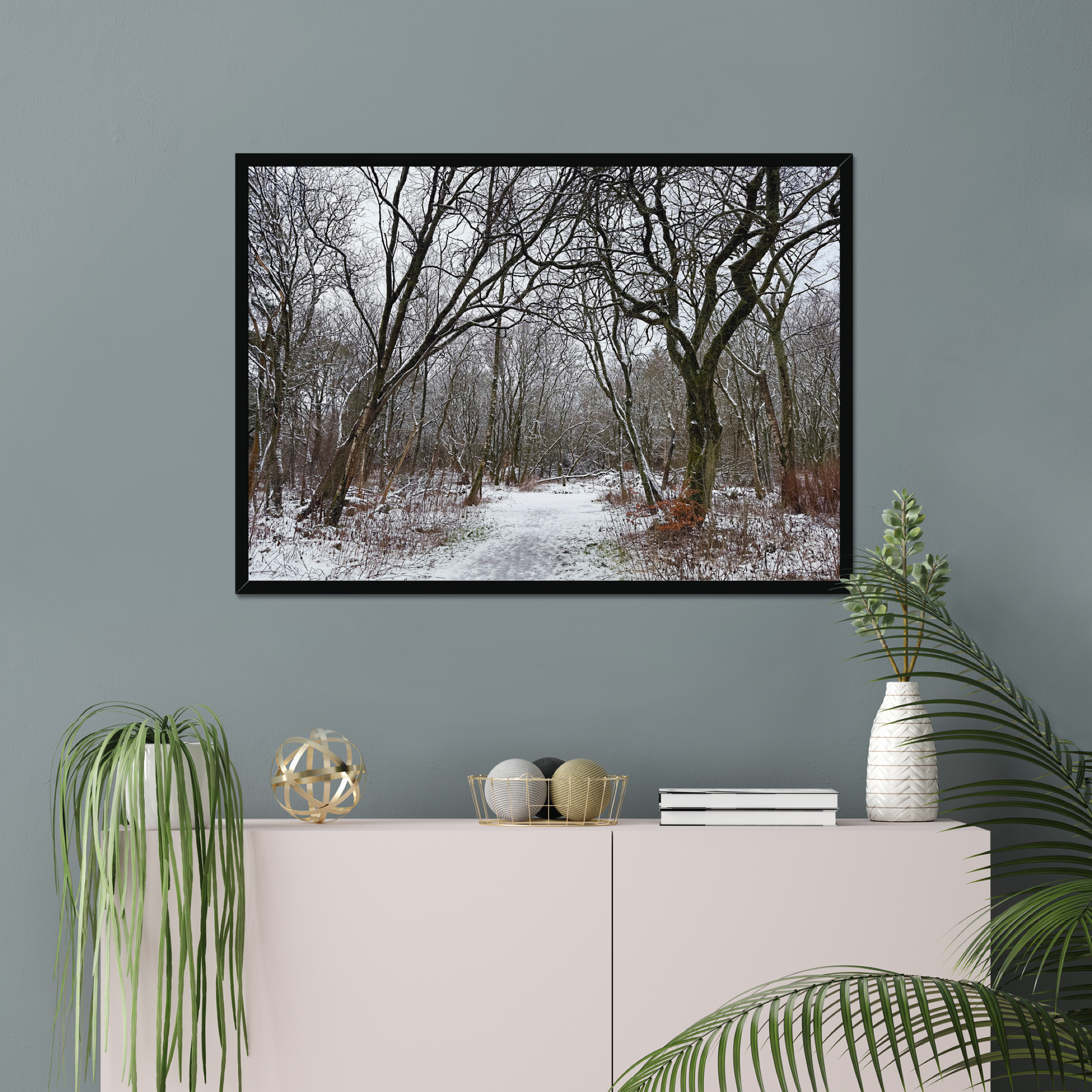 Framed photograph of a winter woodland scene with snow on the ground on a gray wall above a white cabinet with decorative items.