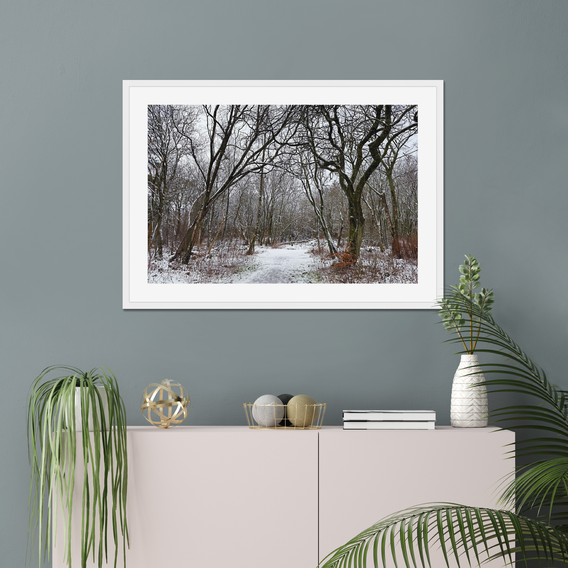 Framed photograph of a winter woodland scene with snow on the ground on a gray wall above a white cabinet with decorative items.