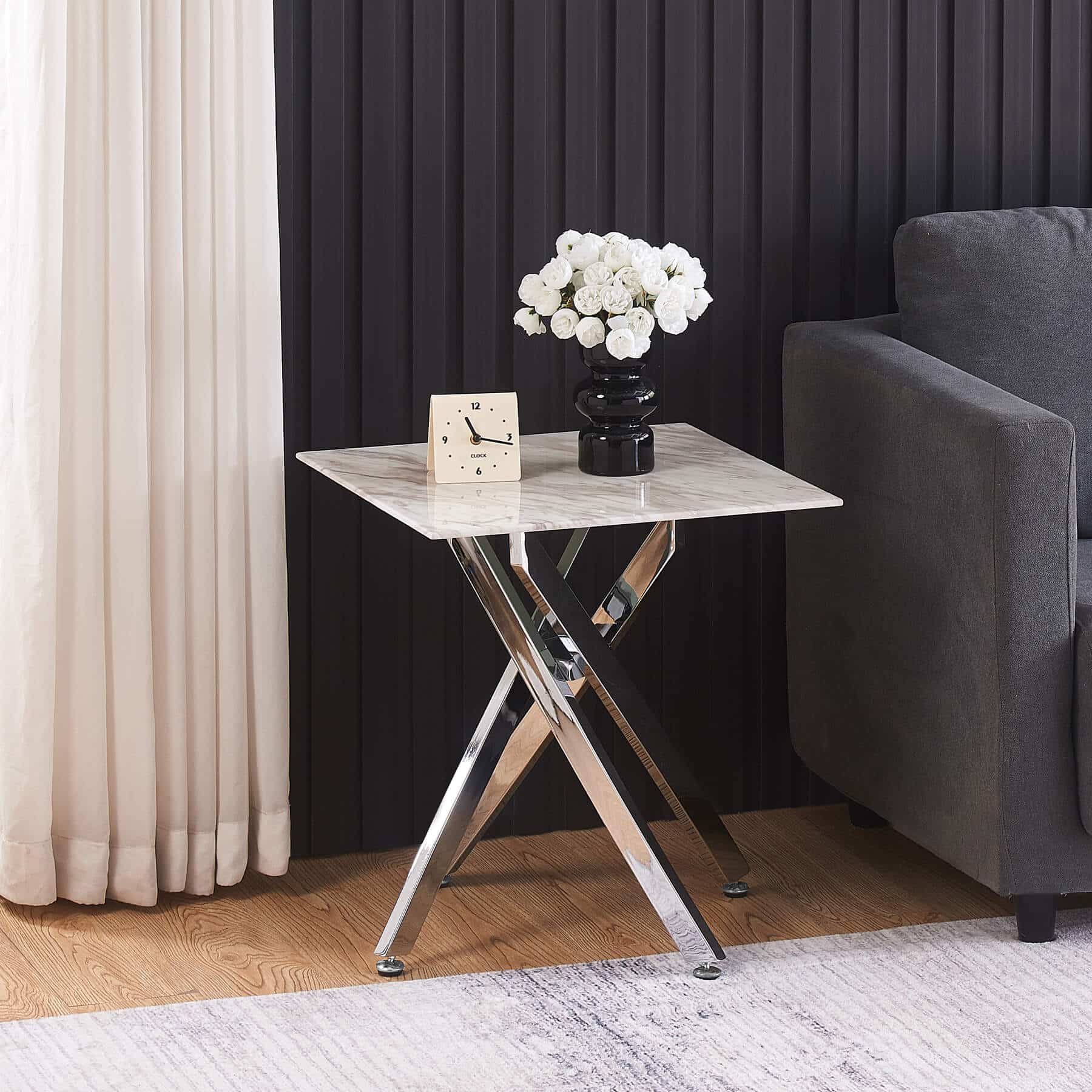 Small marble table with a clock and flower vase next to a gray sofa in a room with black curtains.