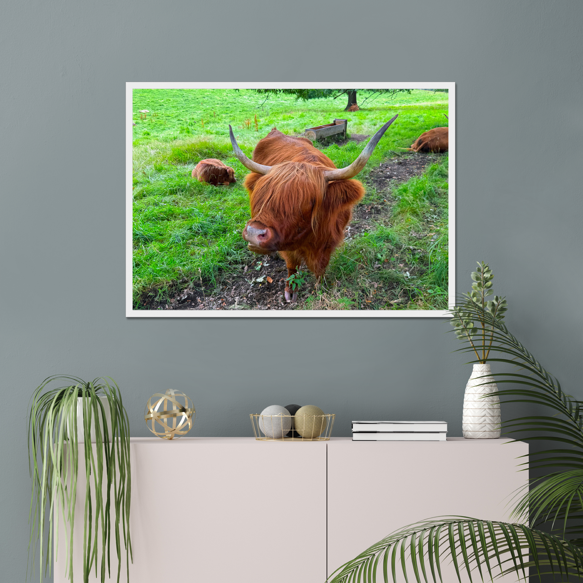 Framed picture of a highland cow on a wall above a cabinet with decorative items.