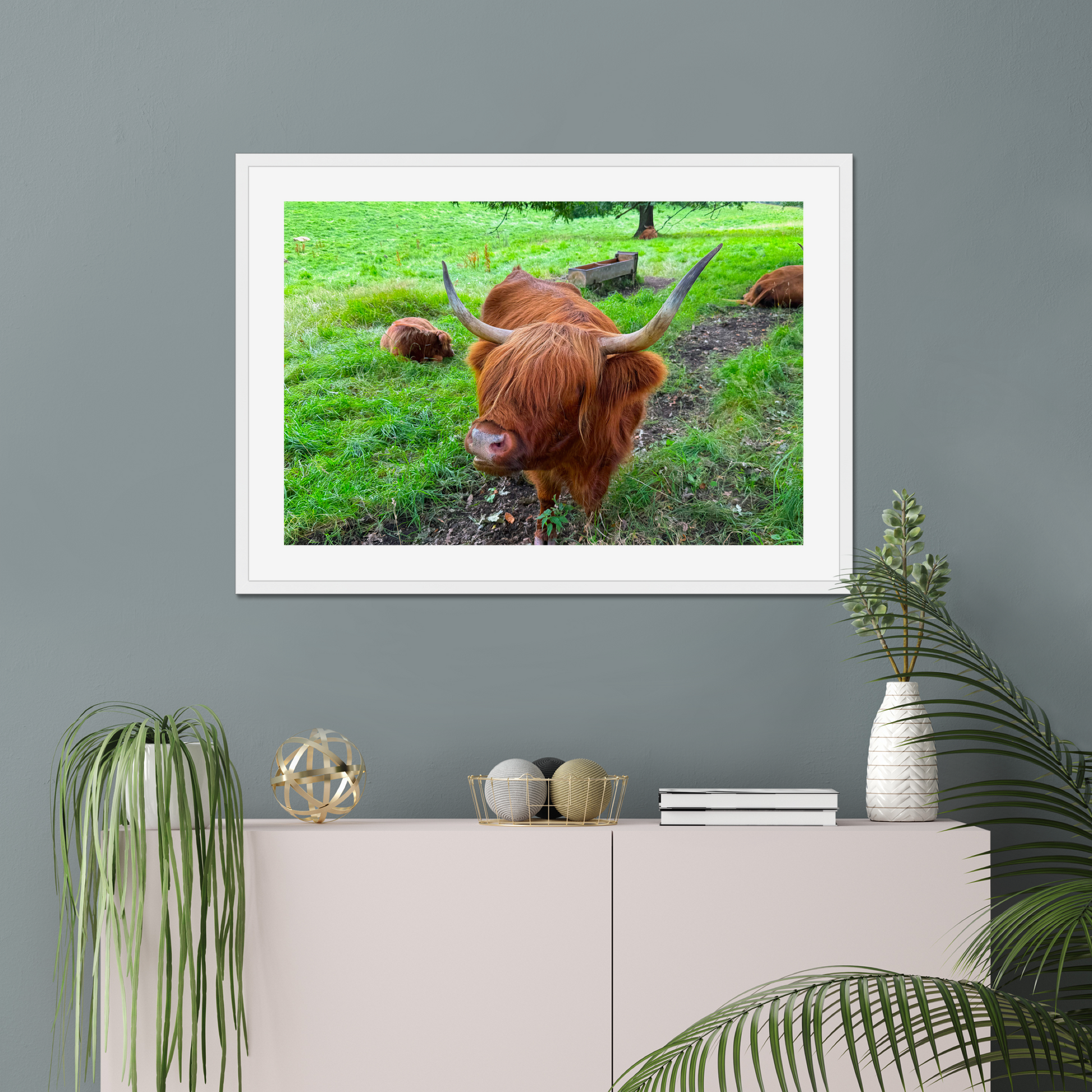 Framed picture of a highland cow on a wall above a cabinet with decorative items.