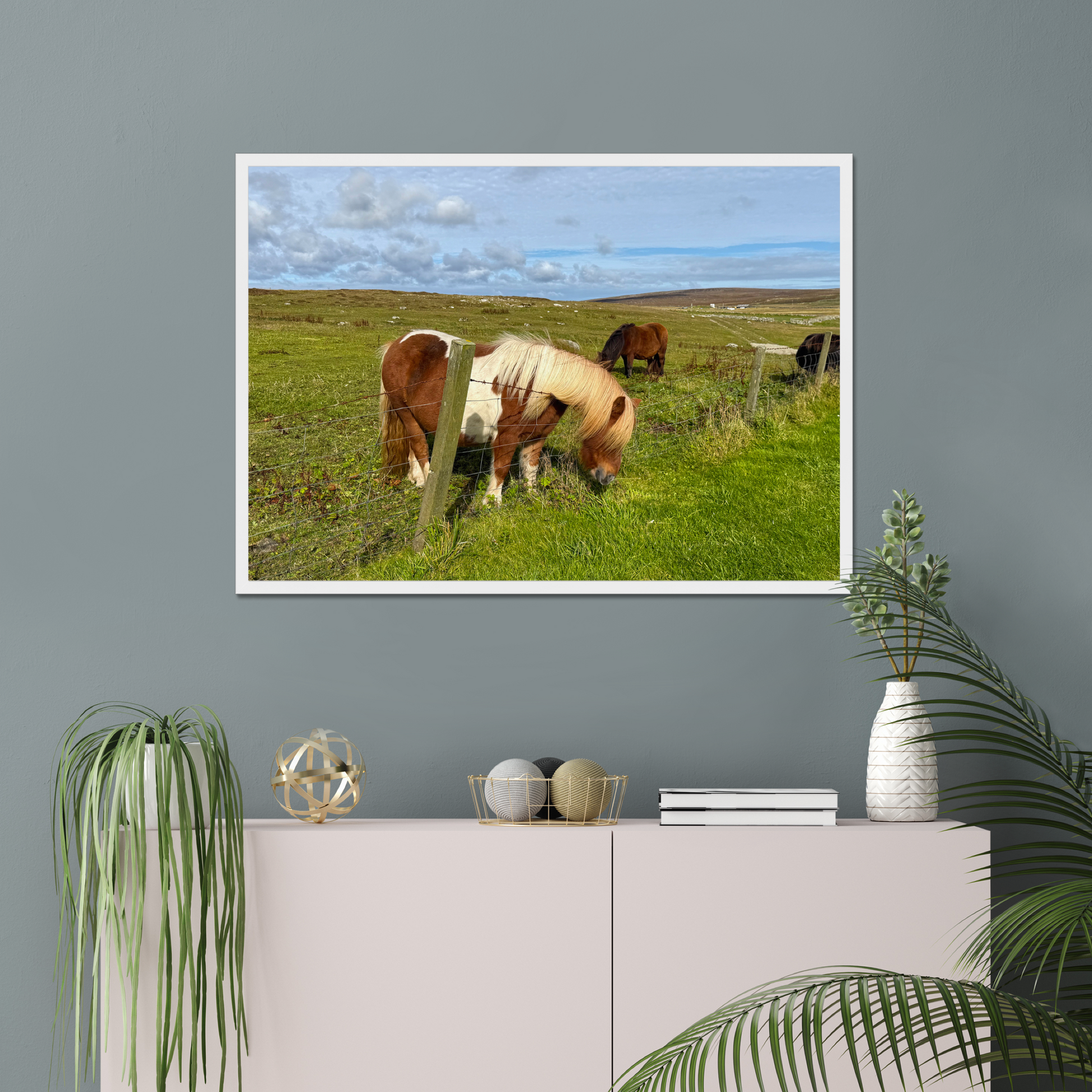 Framed picture of horses grazing on a grassy field hanging on a gray wall above a white cabinet with decorative items.