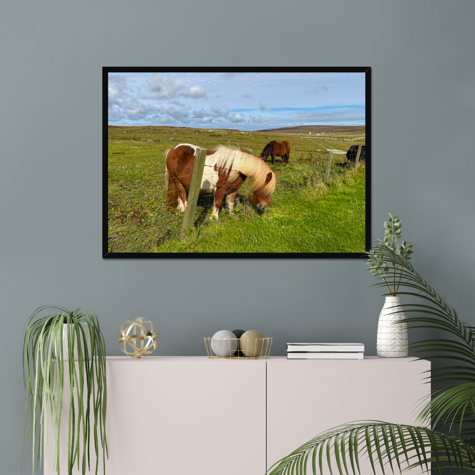 Framed picture of horses in a field on a gray wall above a white cabinet with decorative items.