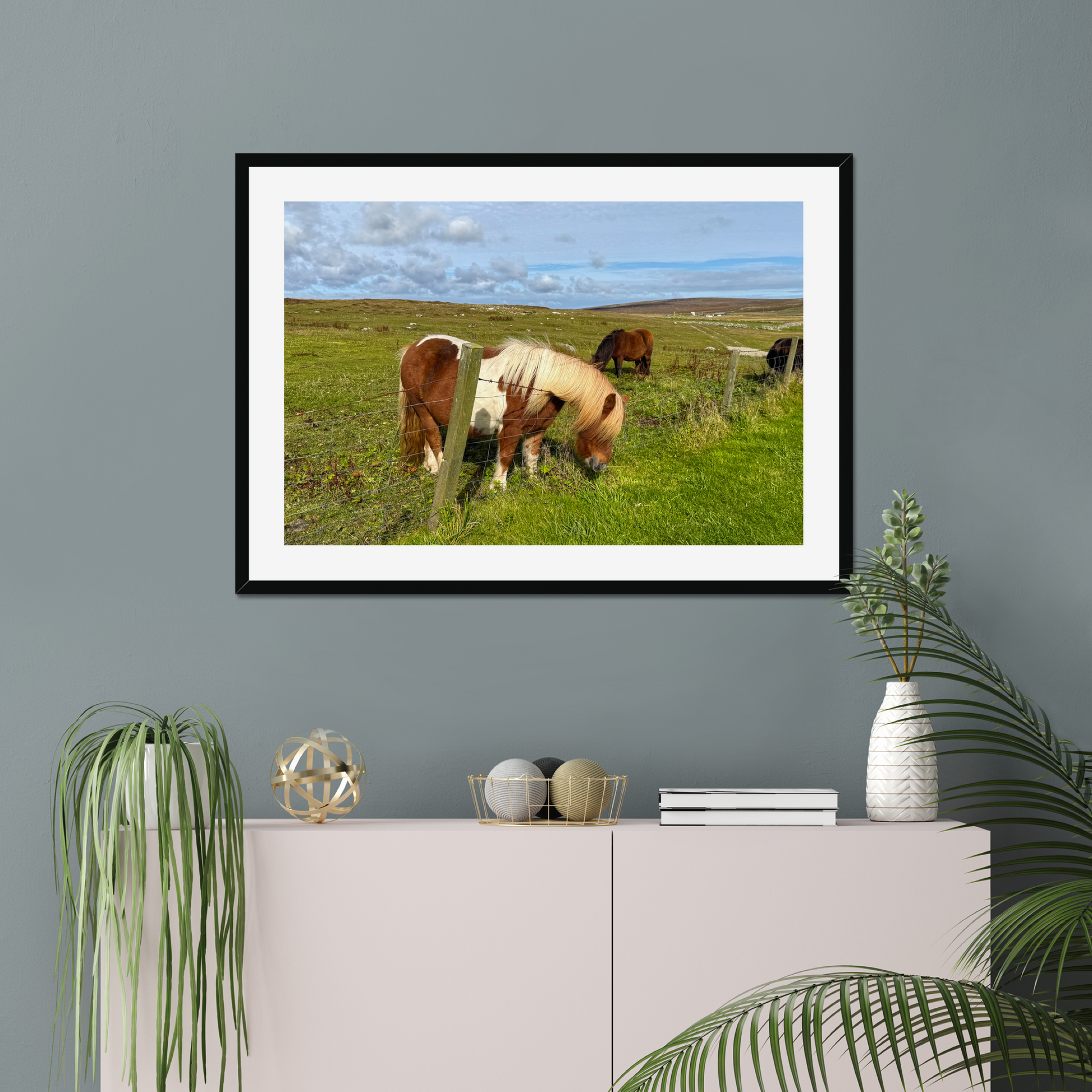 Framed picture of horses grazing on a grassy field hanging on a gray wall above a white cabinet with decorative items.