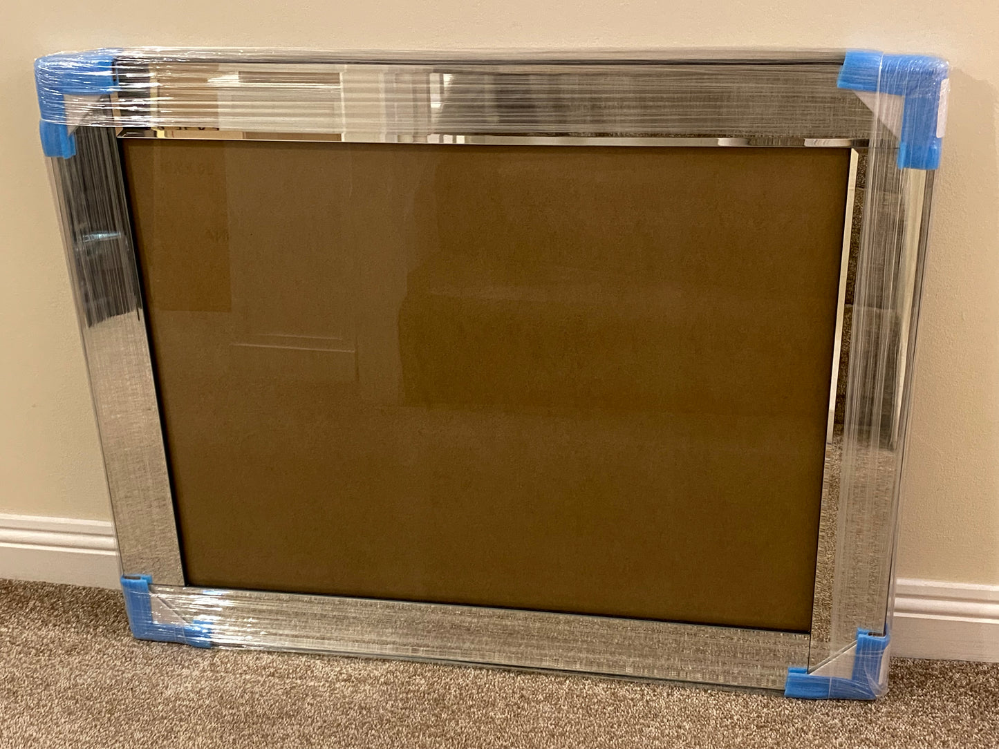 Mirrored picture frame with clear plastic wrap and blue tape on a beige wall and carpeted floor.