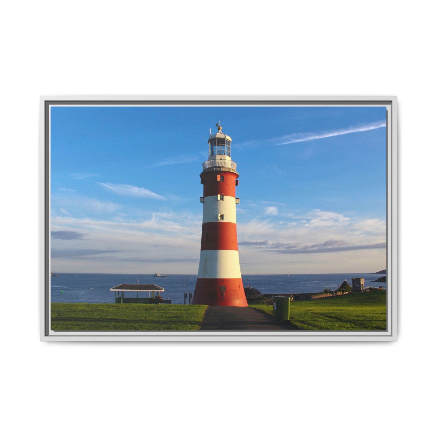 Red and white lighthouse on a grassy area with a blue sky and ocean in the background.