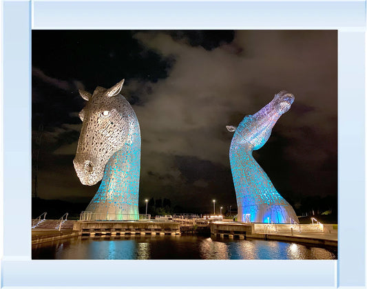 Two large horse head sculptures illuminated in blue and green by a body of water at night.