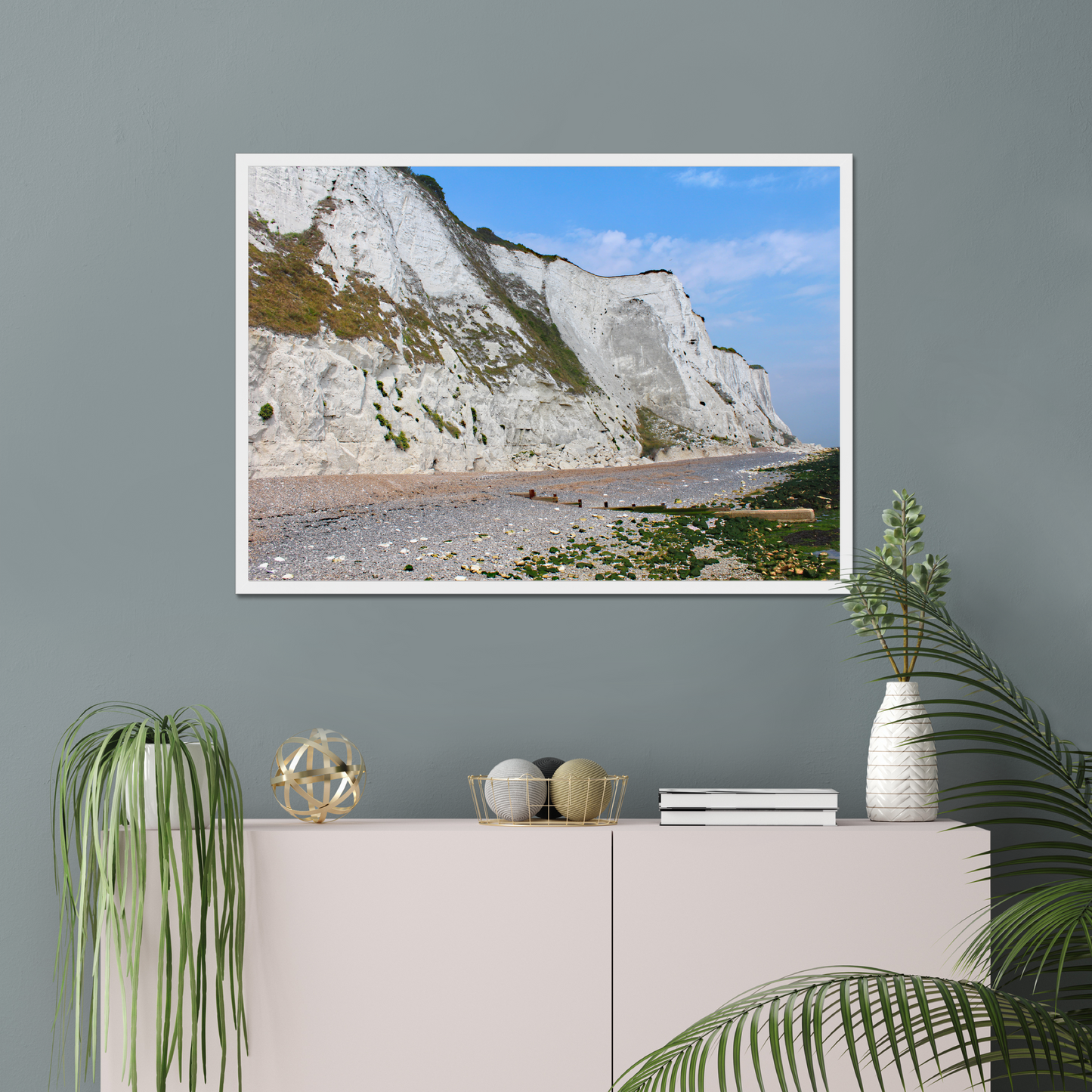 Framed photo of white cliffs with a pebbly beach and blue sky on a gray wall above a white cabinet with decorative items.