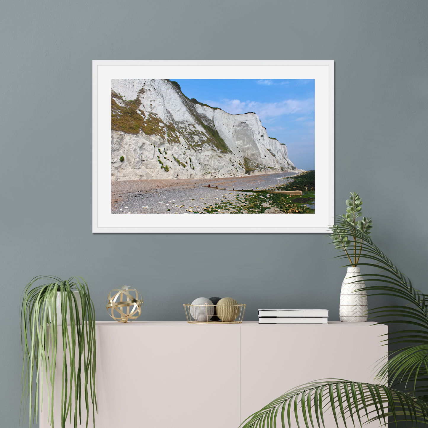 Framed photo of white cliffs with a pebbly beach and blue sky on a gray wall above a white cabinet with decorative items.
