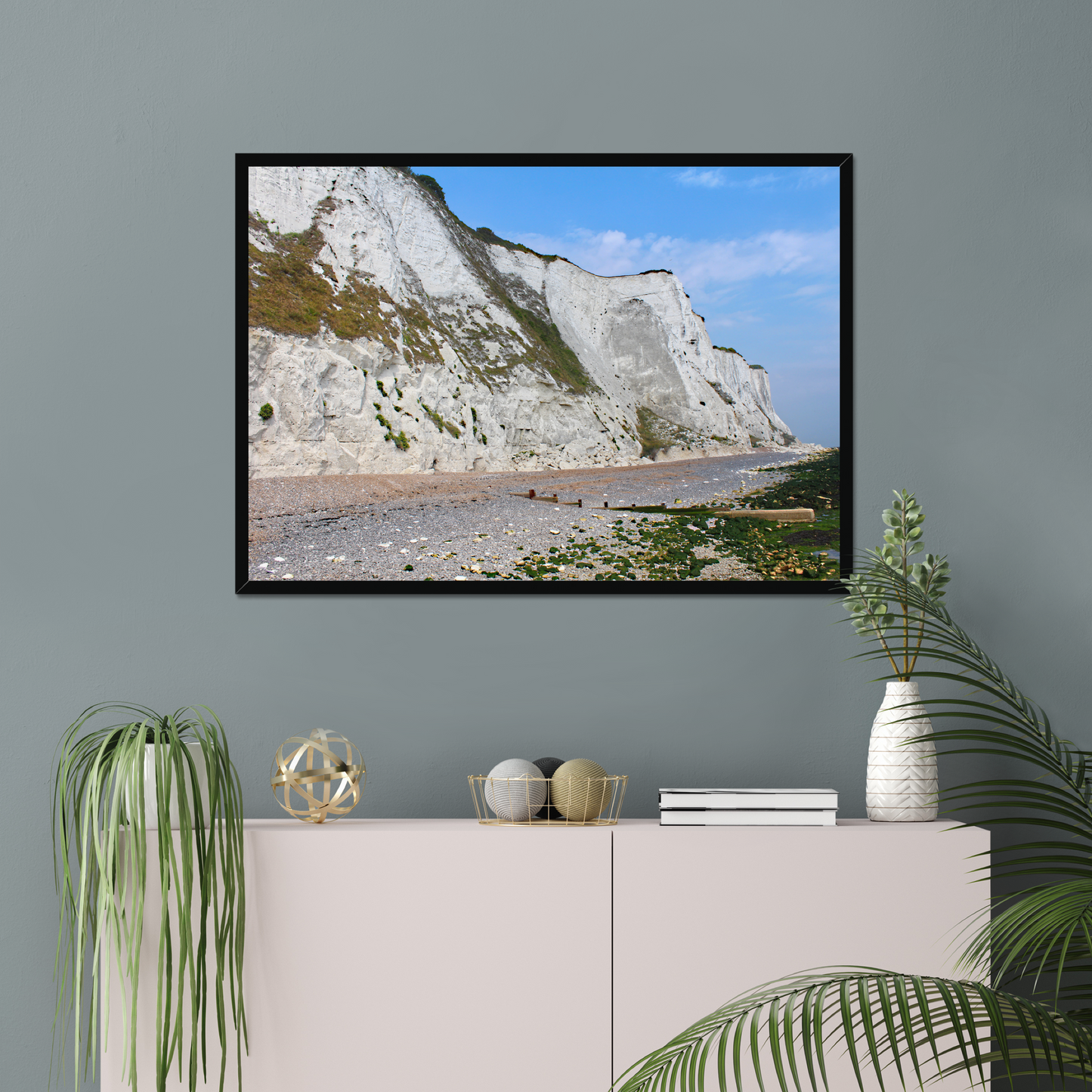 Framed photo of white cliffs with a pebbly beach and blue sky on a gray wall above a white cabinet with decorative items.