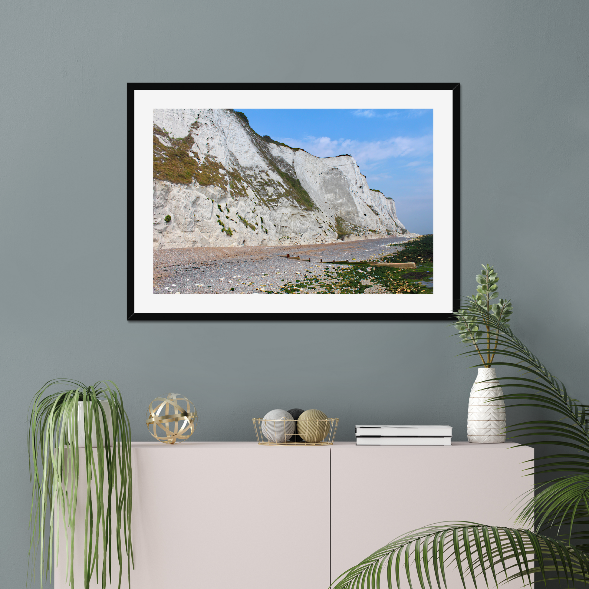 Framed photo of white cliffs with a pebbly beach and blue sky on a gray wall above a white cabinet with decorative items.